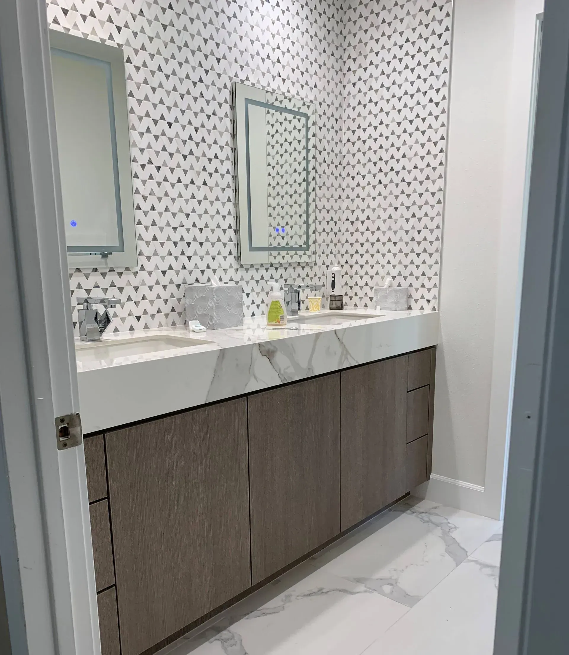 Bathroom with double sink vanity, mirrors, patterned tile backsplash, and wood cabinets.