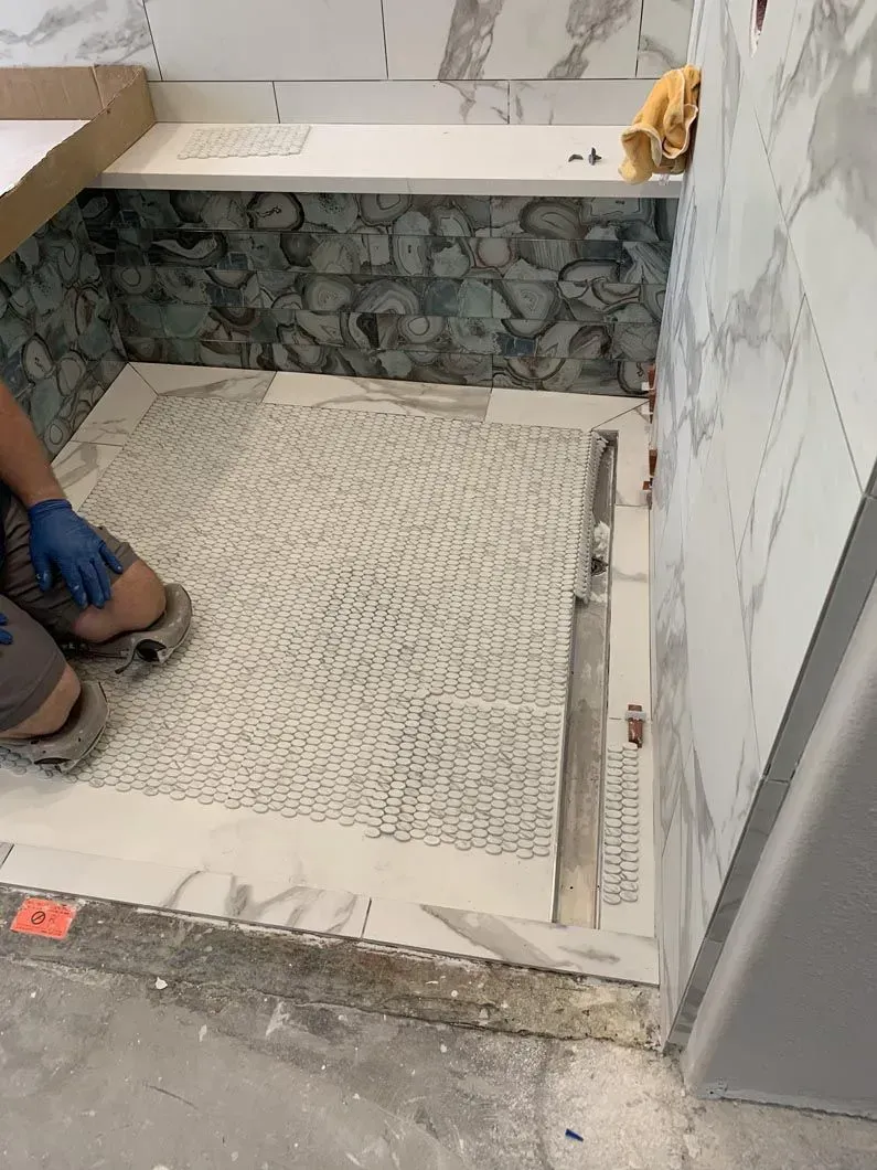 Person installing mosaic tile in a shower. White and gray tiles, with blue gloves and gray boots.