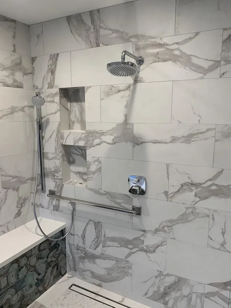 A marble-tiled shower with a bench, hand shower, overhead shower, grab bar, and recessed storage niche.