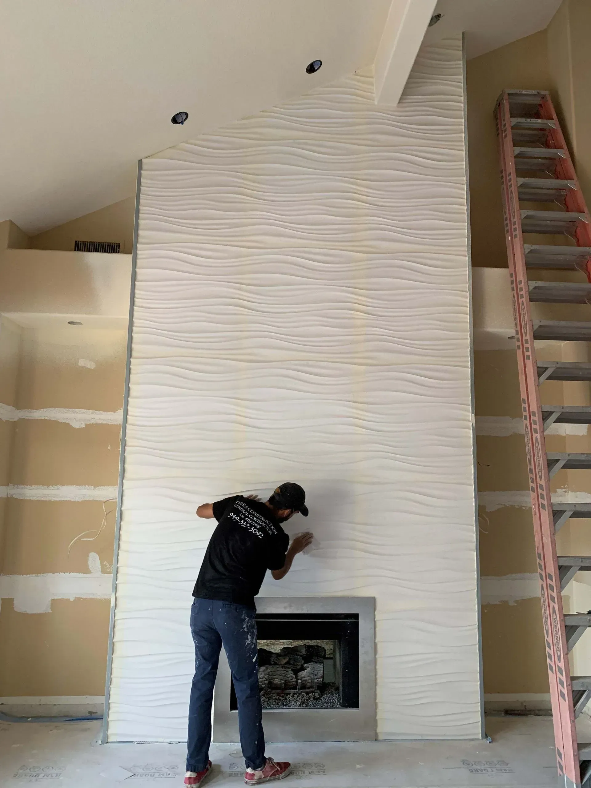 Man working on white textured fireplace in a room with a ladder, trim, and a fireplace.