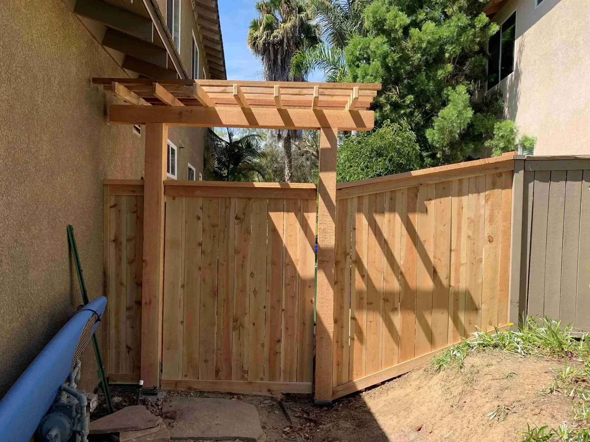 Wooden fence with a gate and pergola at the entrance.  The gate is open and the sky is blue.