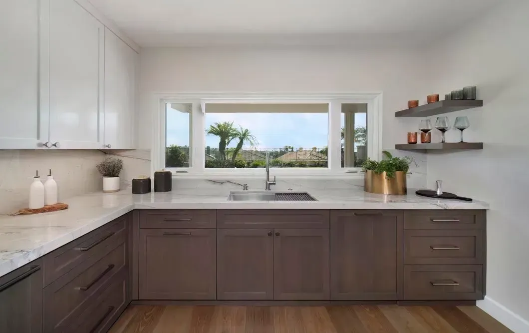 Modern kitchen with white and brown cabinets, window over sink, shelves with glasses and bottles.