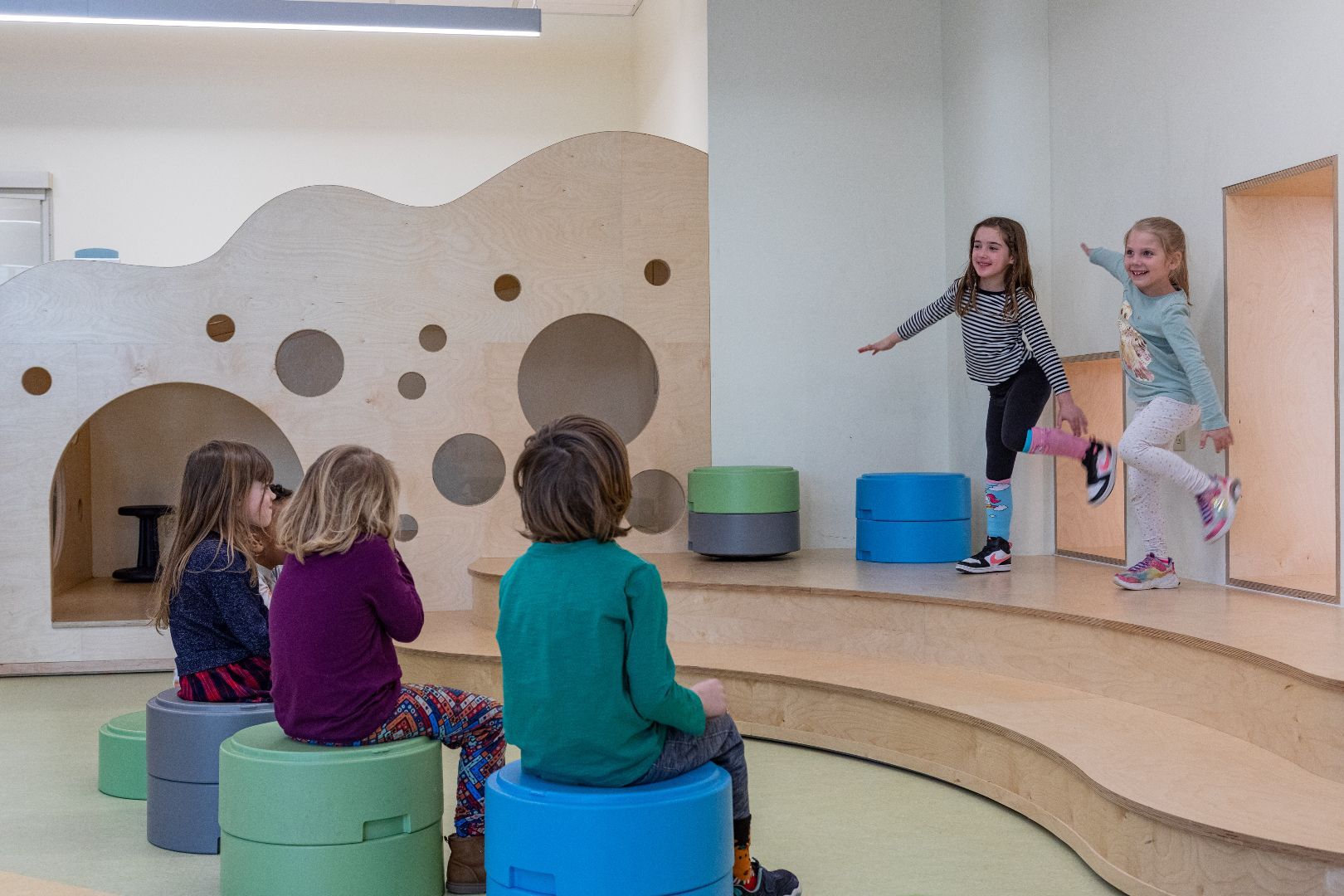 A group of children are sitting on blue ottomans in a room