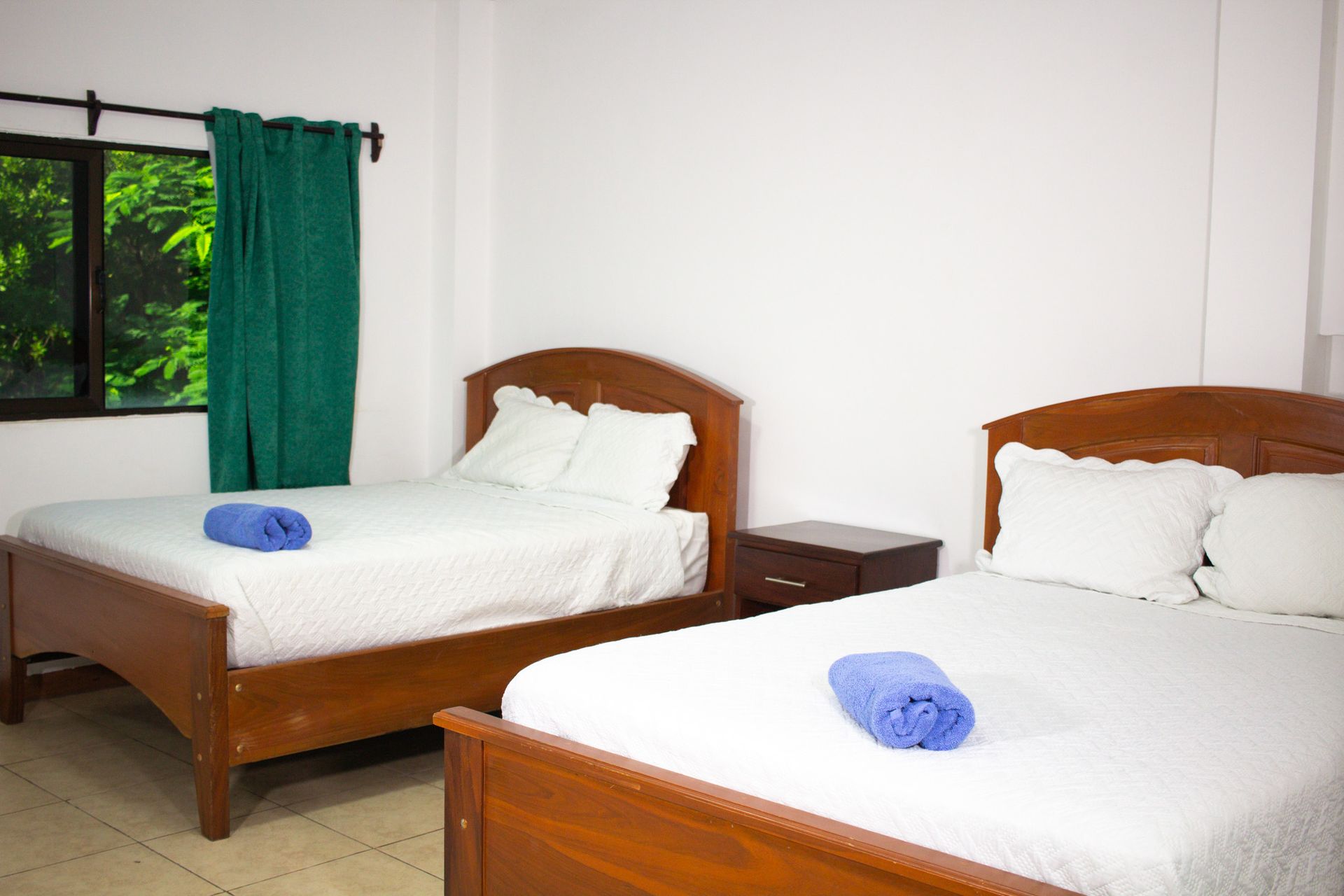 Bedroom with two wooden beds, white linens, blue towels. Green curtains by a window.