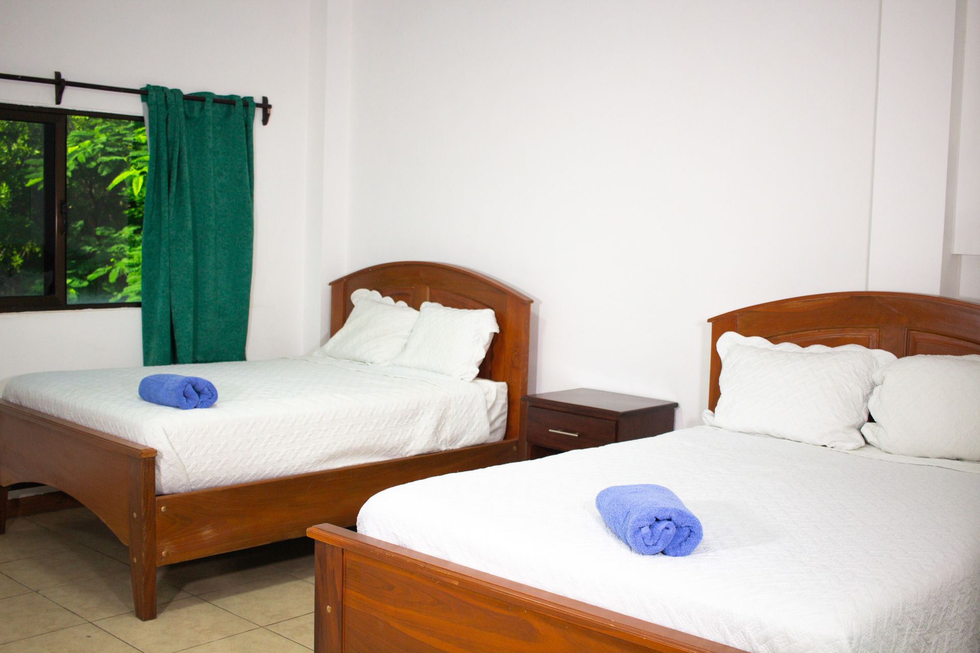 Two beds with white linens, blue towels, and a window with green curtains.