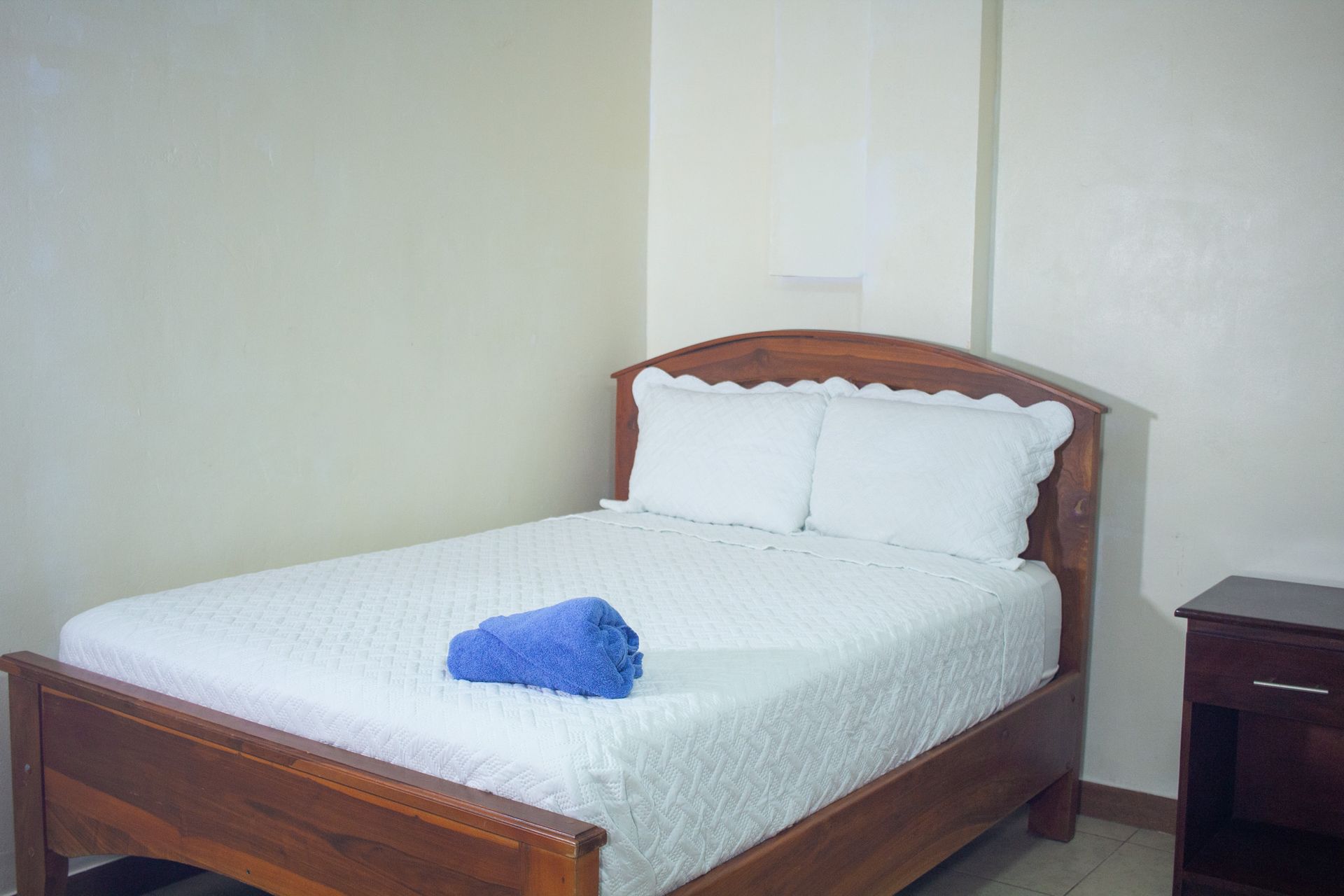 Bedroom with a bed, blue towel, and nightstand against a light-colored wall.