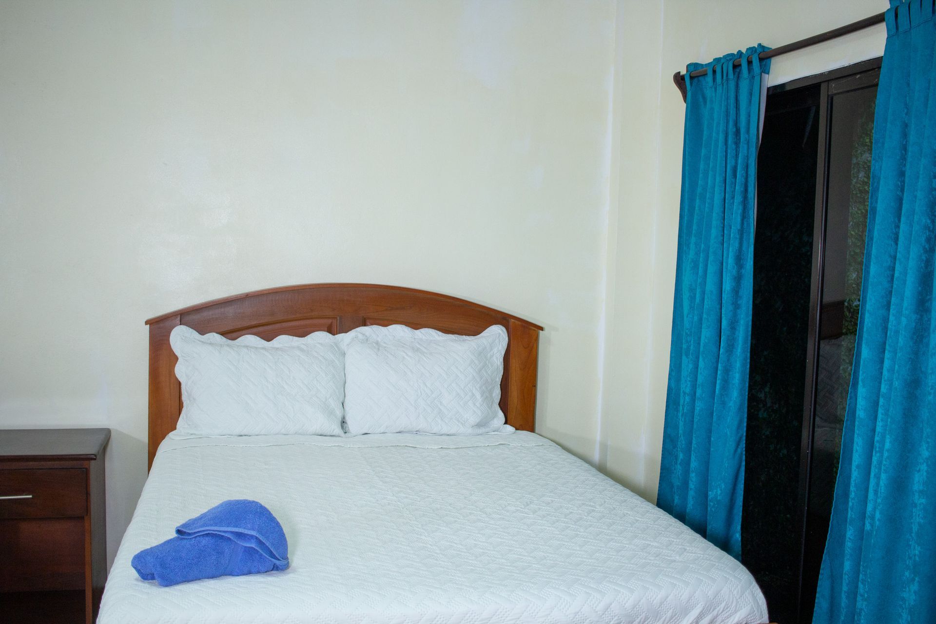Bedroom with a white bed, wooden headboard, turquoise curtains, and a blue towel.