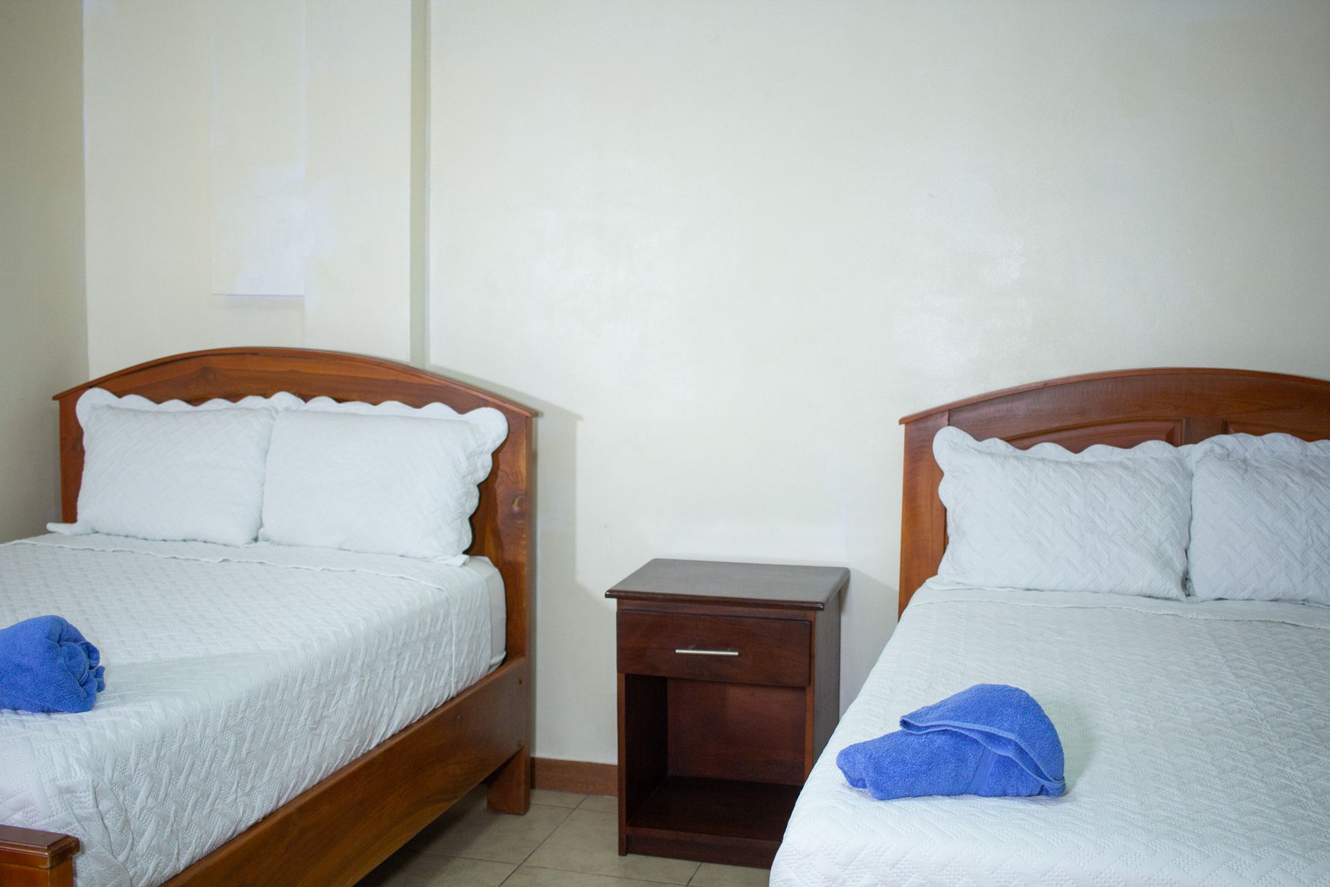 Two beds with white bedding and blue towels in a simple room with a nightstand.
