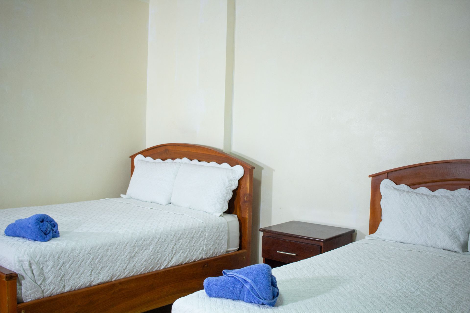 Two beds in a cream-walled room with wooden frames, white linens, and blue towels; a nightstand sits between them.