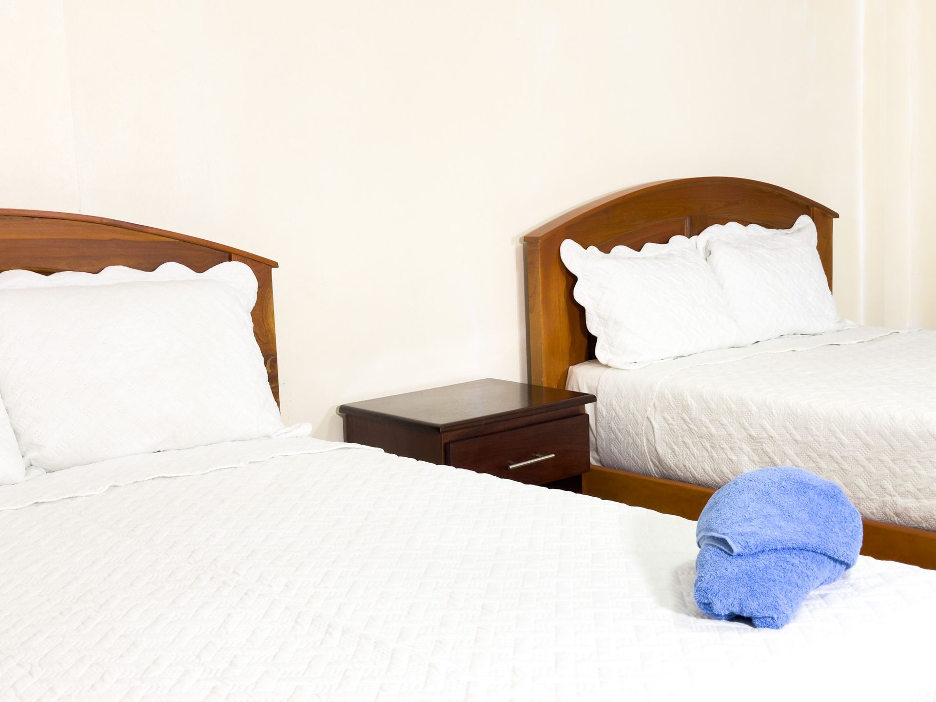 Two beds with white linens and wooden headboards, a nightstand, and a folded blue towel.