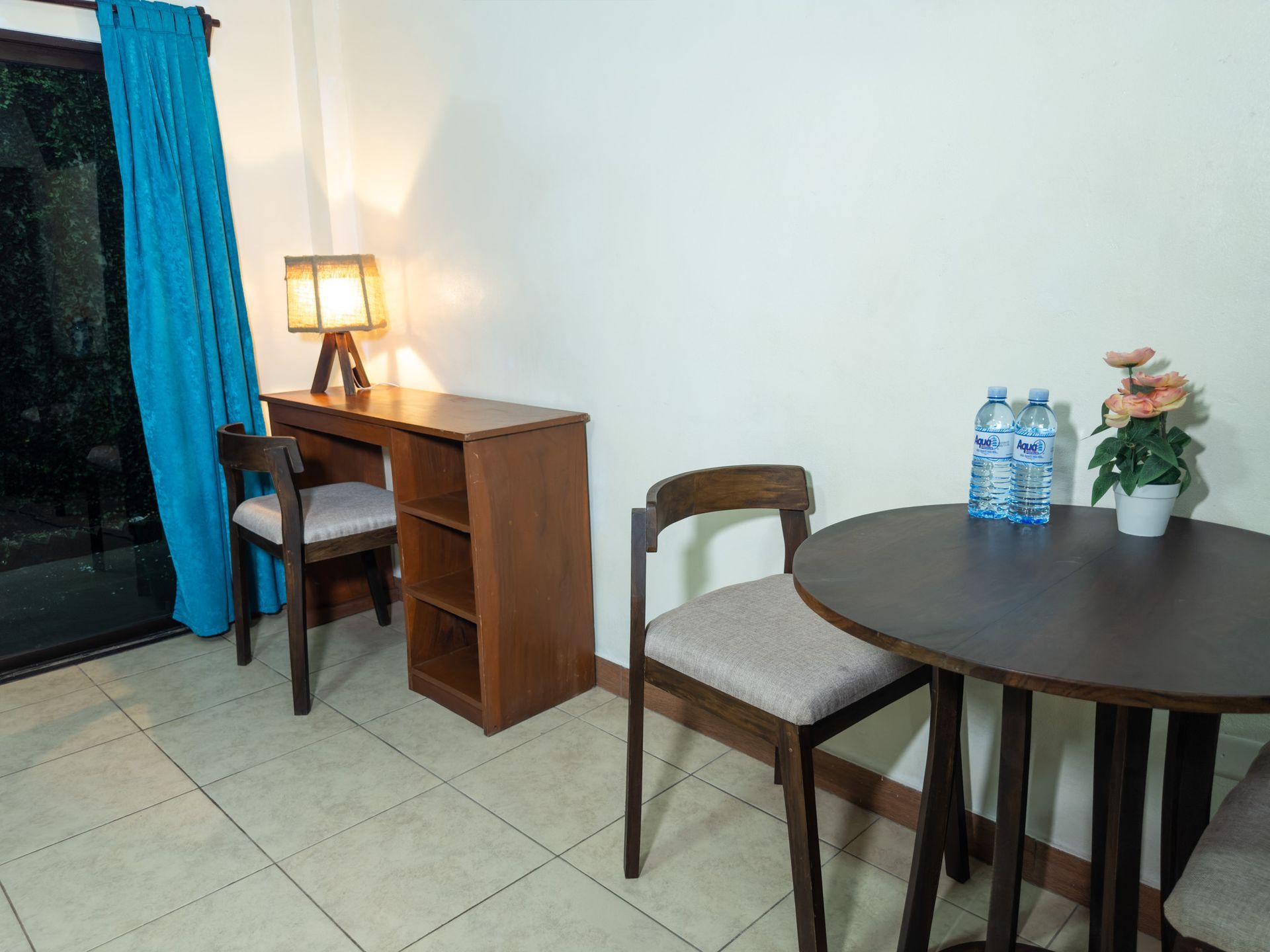 A simple room with a desk, chair, small table, chairs, and flowers. Blue curtain, bright lighting.