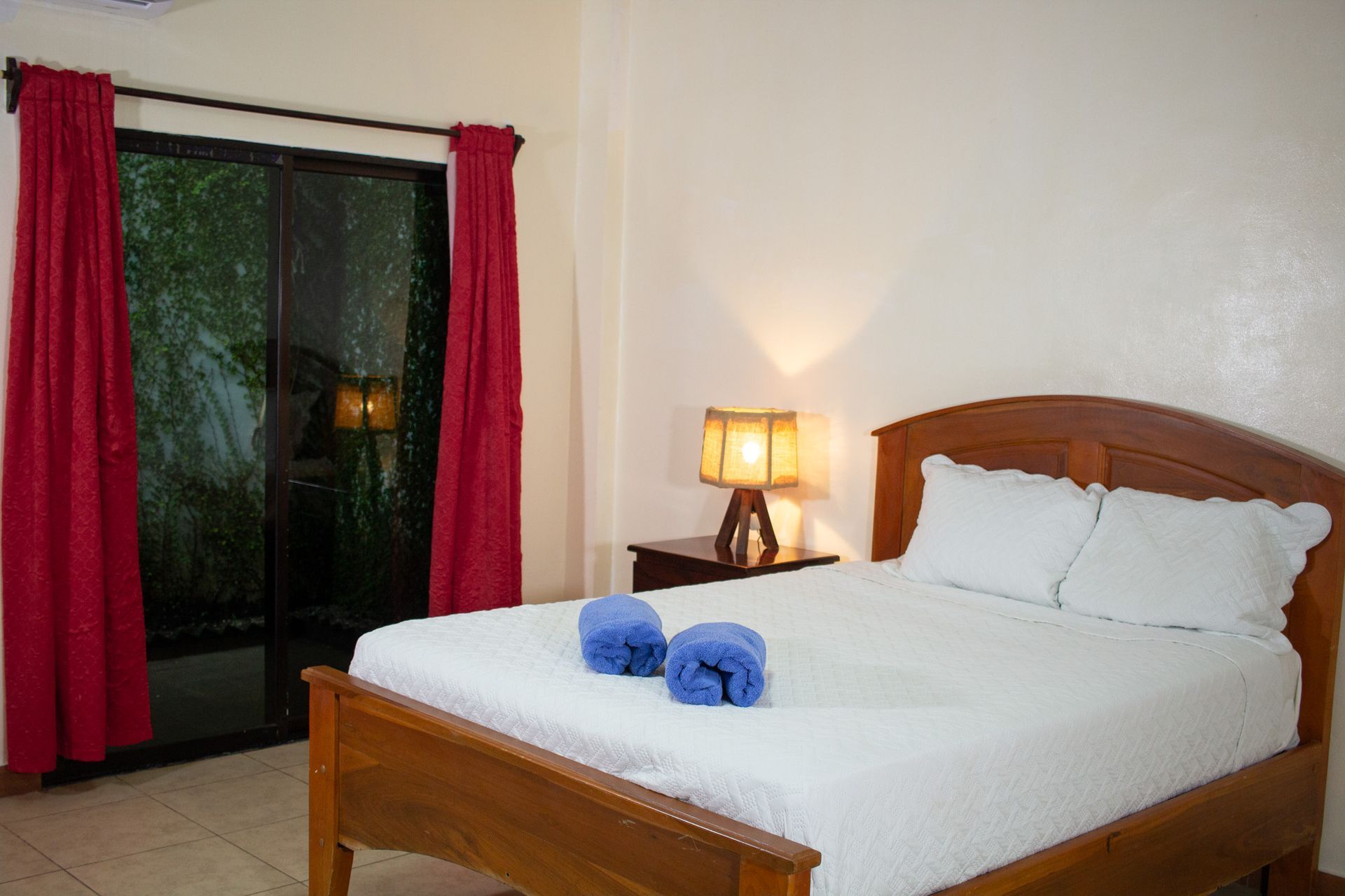 Bedroom with double bed, nightstand, sliding door with red curtains, and blue rolled towels.