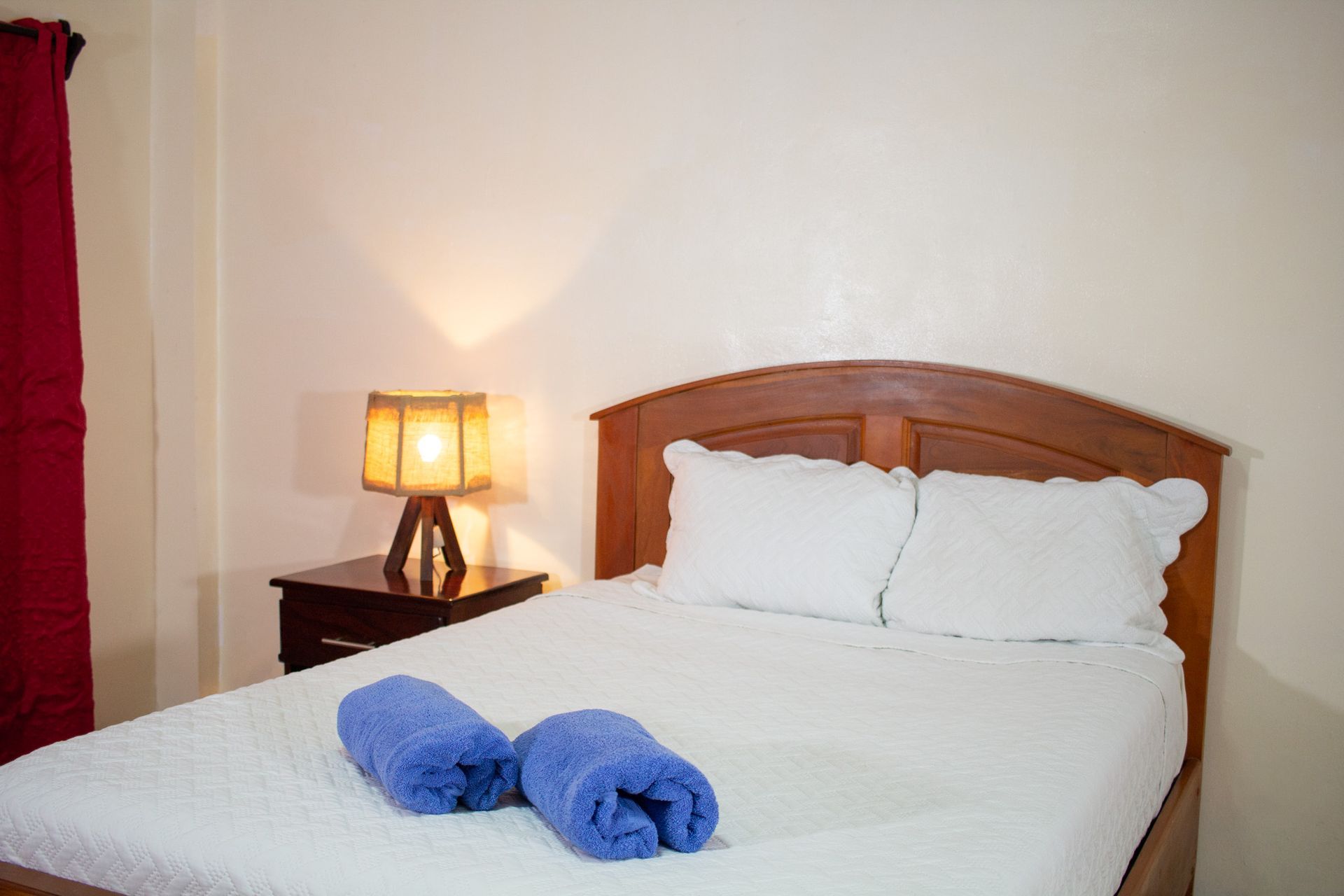 Bedroom with made bed, nightstand, lamp, and rolled blue towels. Red curtain on left.