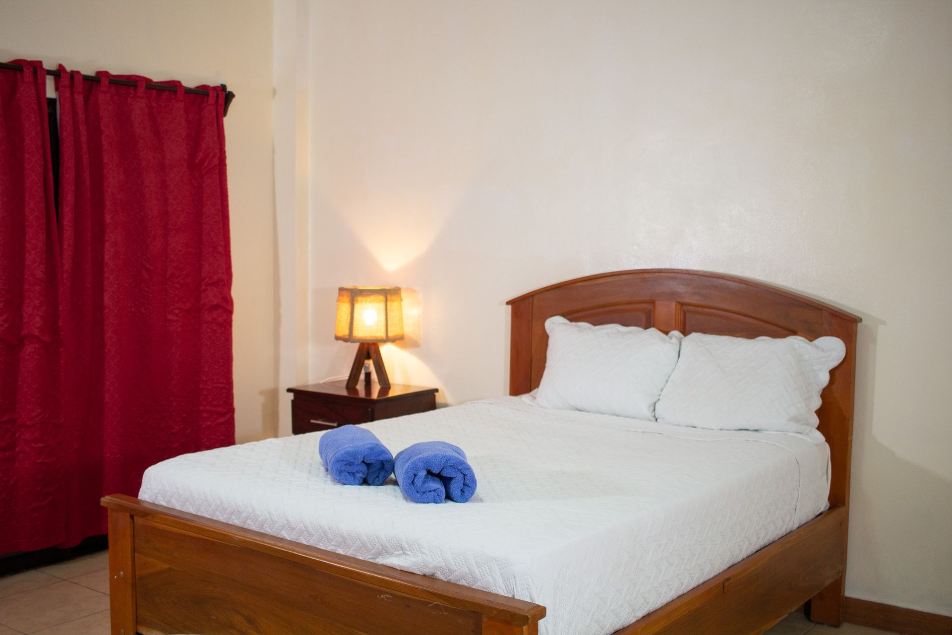 Bedroom with a wooden bed frame, white linens, and a nightstand with a lamp. Red curtains.