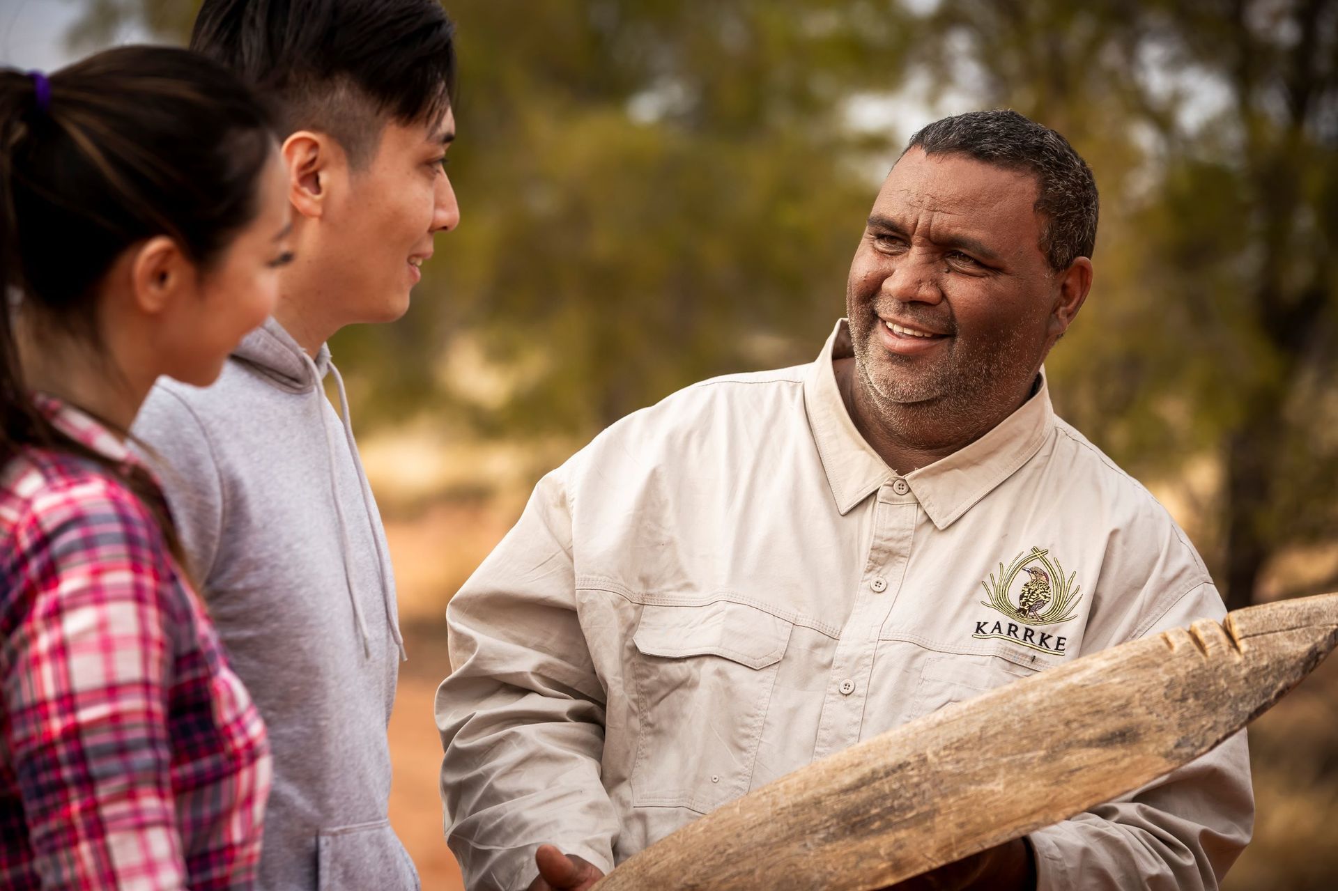 A man is holding a piece of wood and talking to two people.