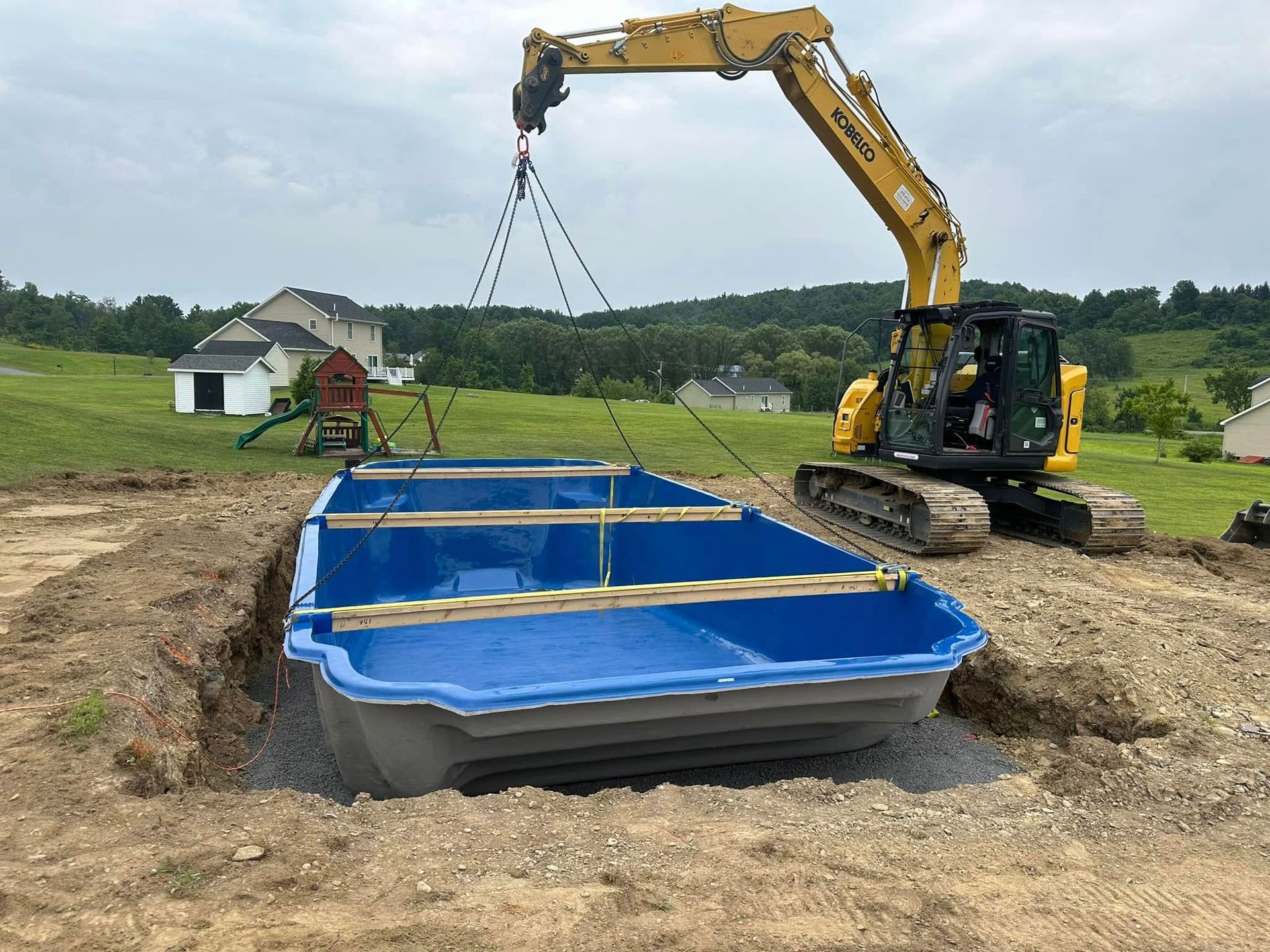An excavator lowers a blue fiberglass pool into an excavated hole in a grassy yard.