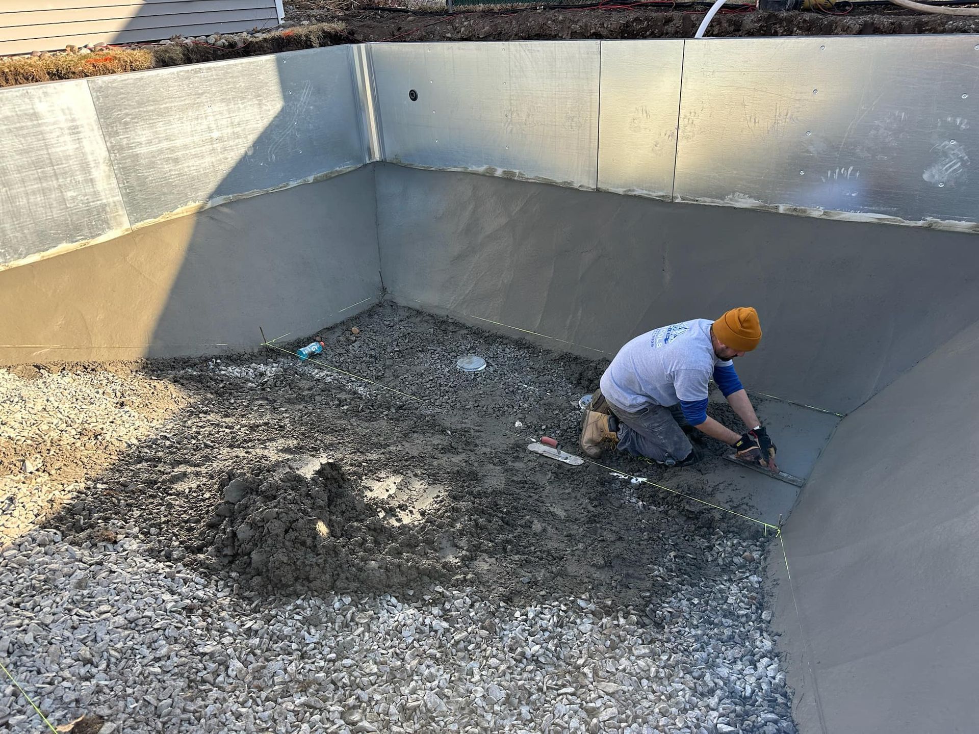Person in a pool shell plastering the wall with a trowel, wearing a yellow beanie, and a gray-walled pool.