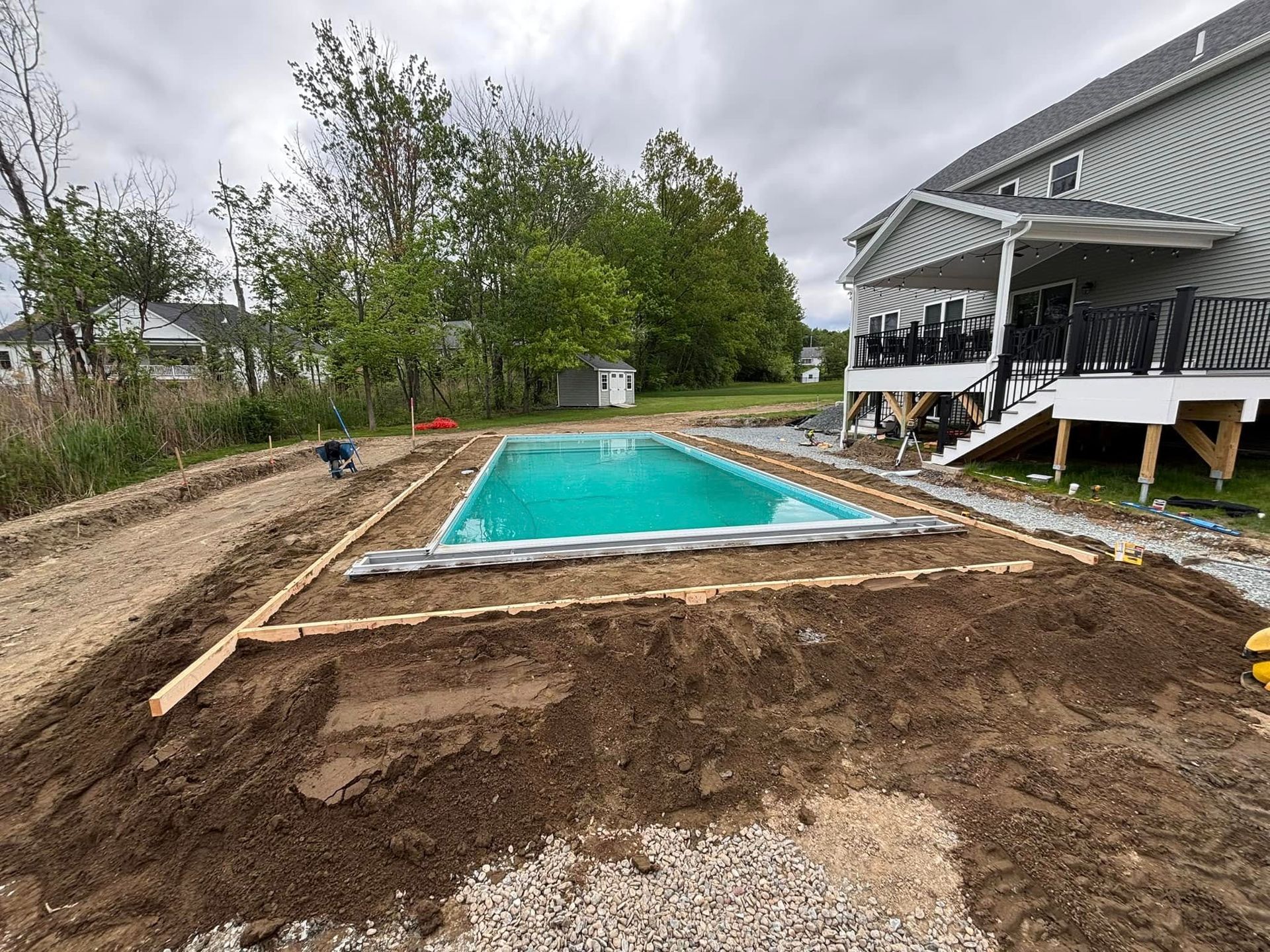 Pool installation in progress next to a house with a deck; earth, gravel, and wooden framing are visible.