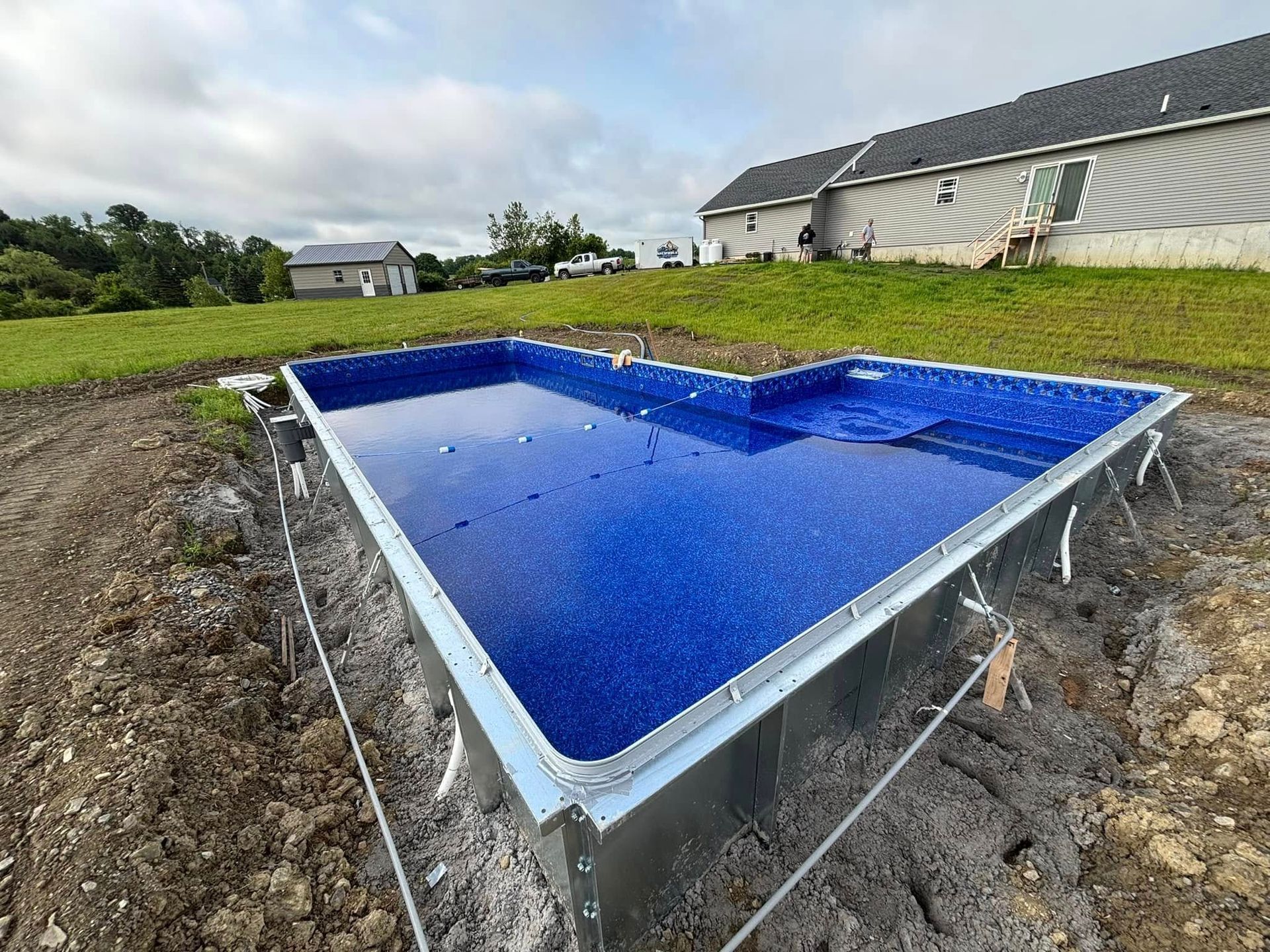 Partially constructed blue tiled pool, surrounded by dirt, near a house and grassy field.