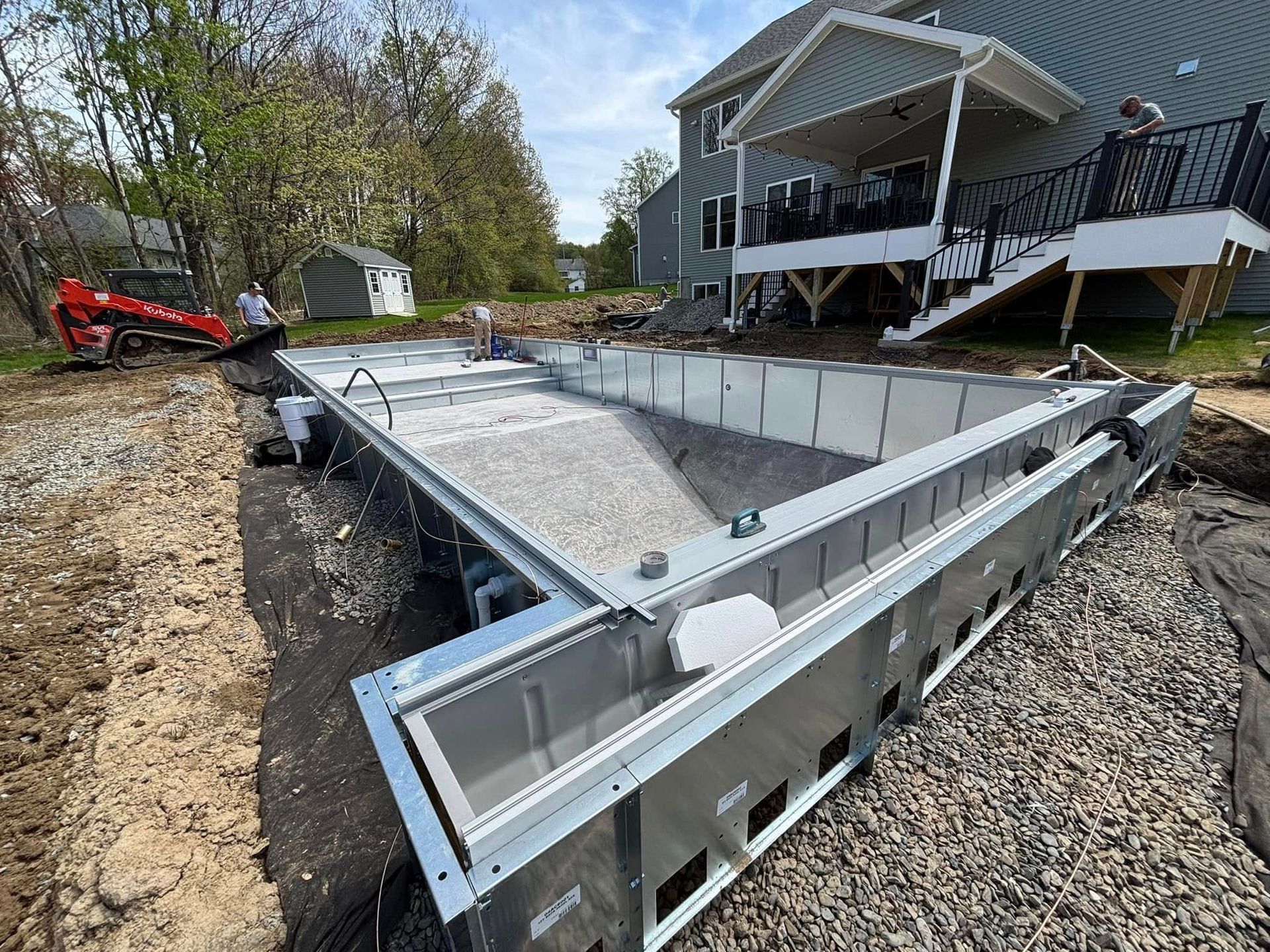 Pool under construction in a yard; metal structure with gravel interior, near a house with a deck.