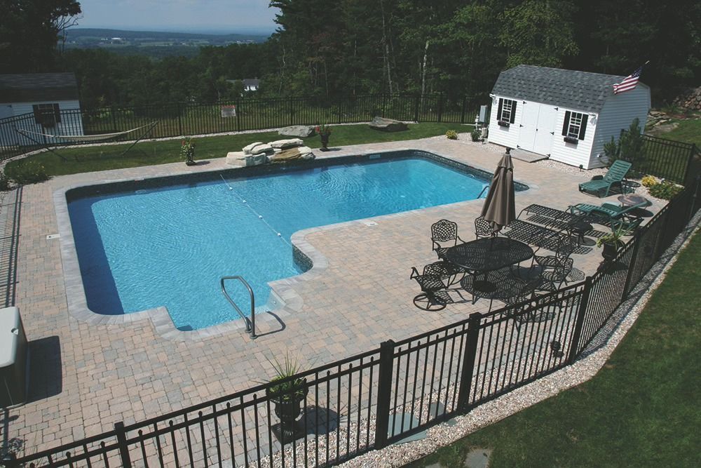 Swimming pool with blue water and surrounding patio, shed, and black fence, with a view of hills in the distance.