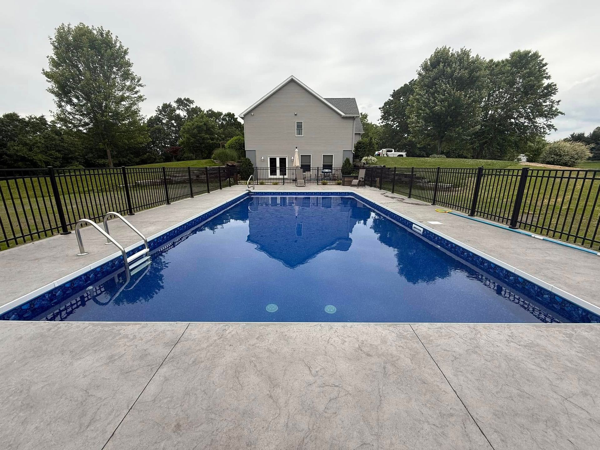 Rectangular blue swimming pool in a backyard, with a house in the background and a black fence.