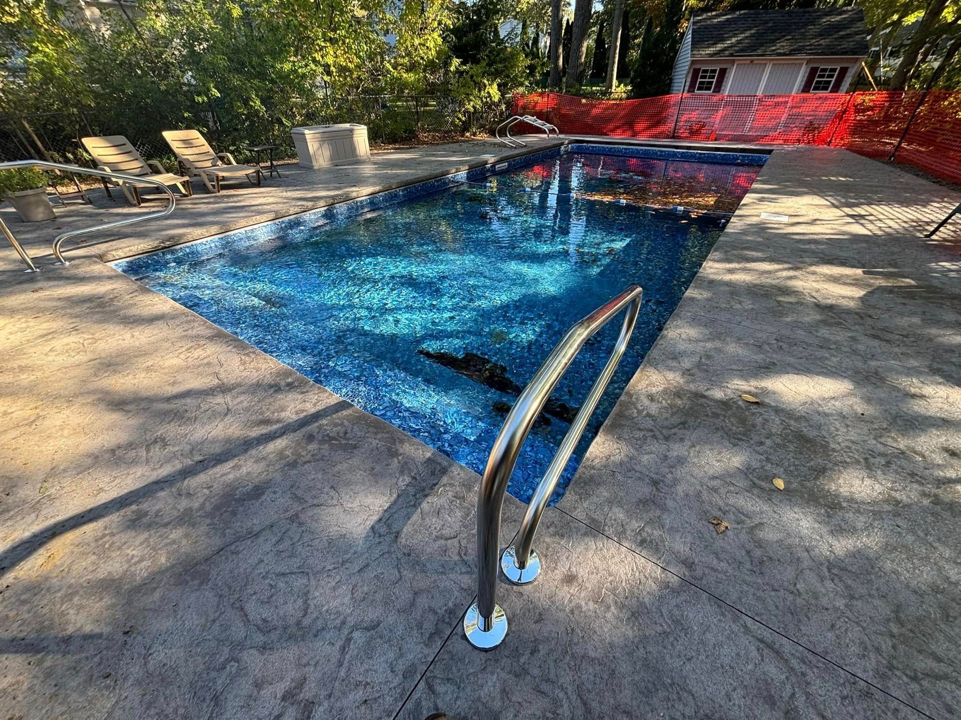 Pool with blue tile, surrounded by concrete deck. Stainless steel ladder. Orange construction fencing in the background.