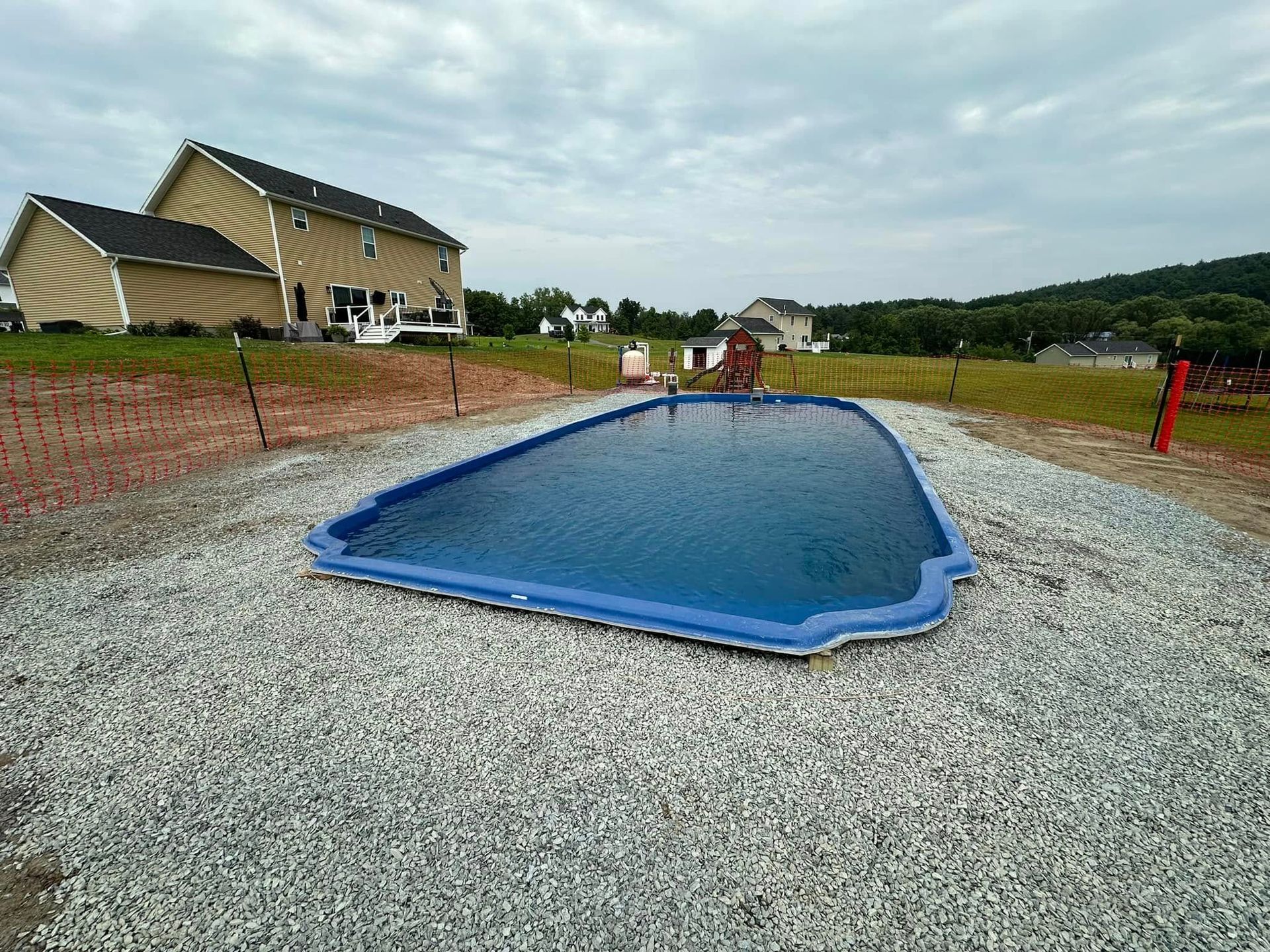 Blue, empty swimming pool in gravel, next to a beige house, under cloudy sky.