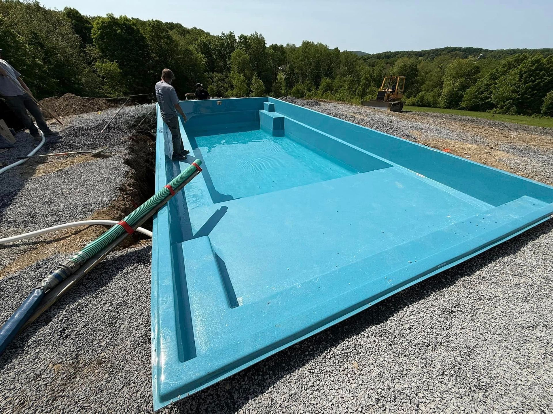 Blue swimming pool under construction, filled with water, near gravel and trees. Two workers near pool.