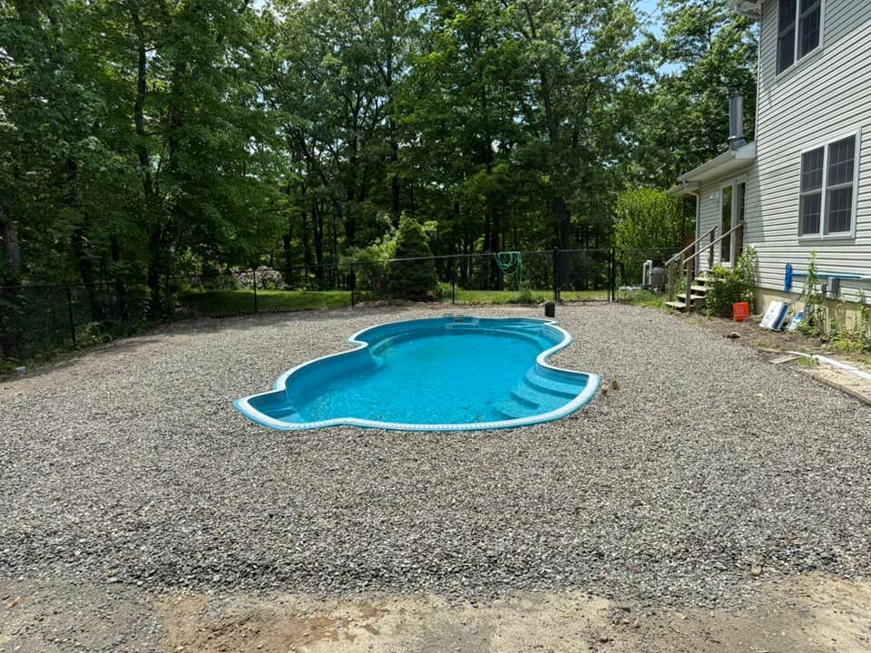 Small turquoise pool surrounded by gray gravel in a backyard, near a white house and trees.