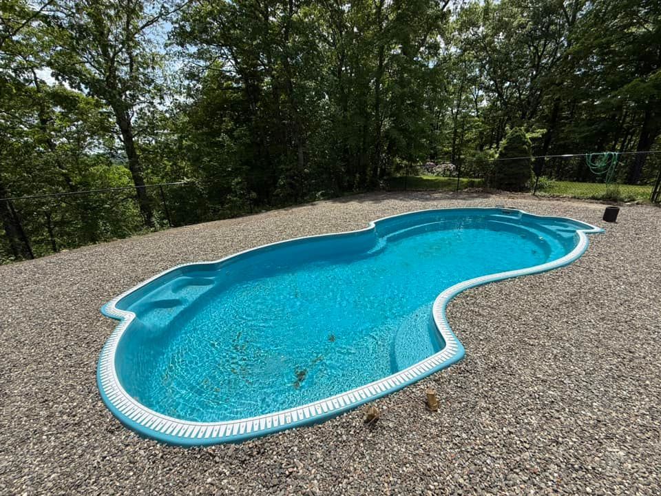 Blue swimming pool with white trim, on gravel, surrounded by trees.