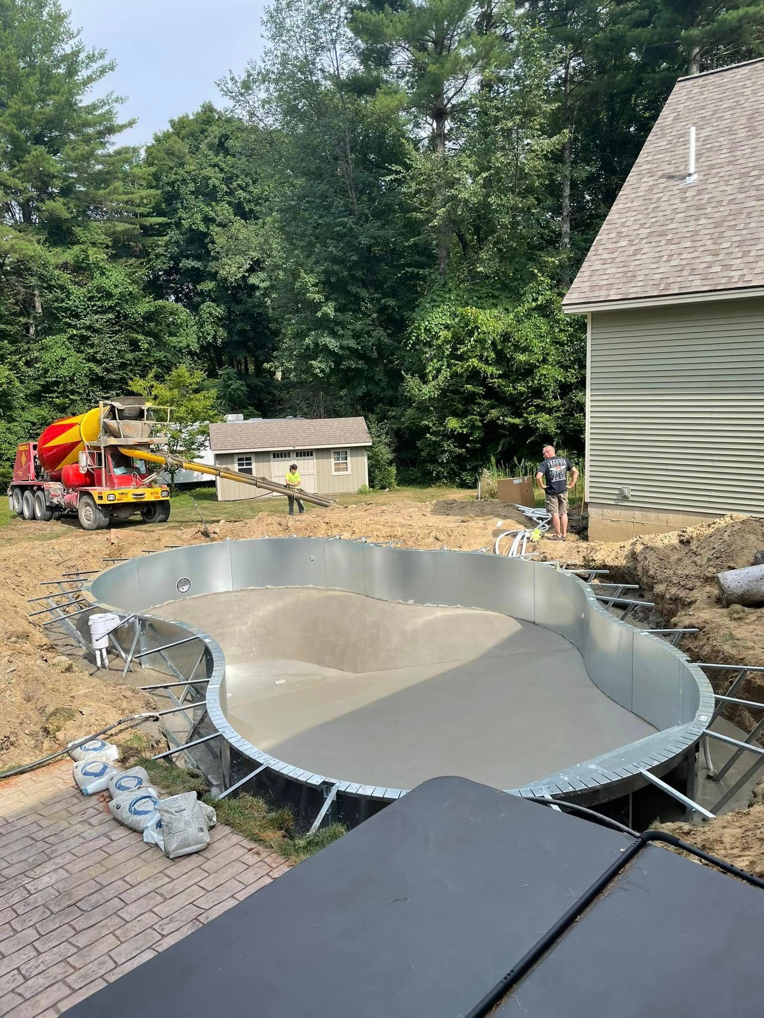 Pool under construction; concrete being poured into metal frame. Construction site with truck, trees, and house.