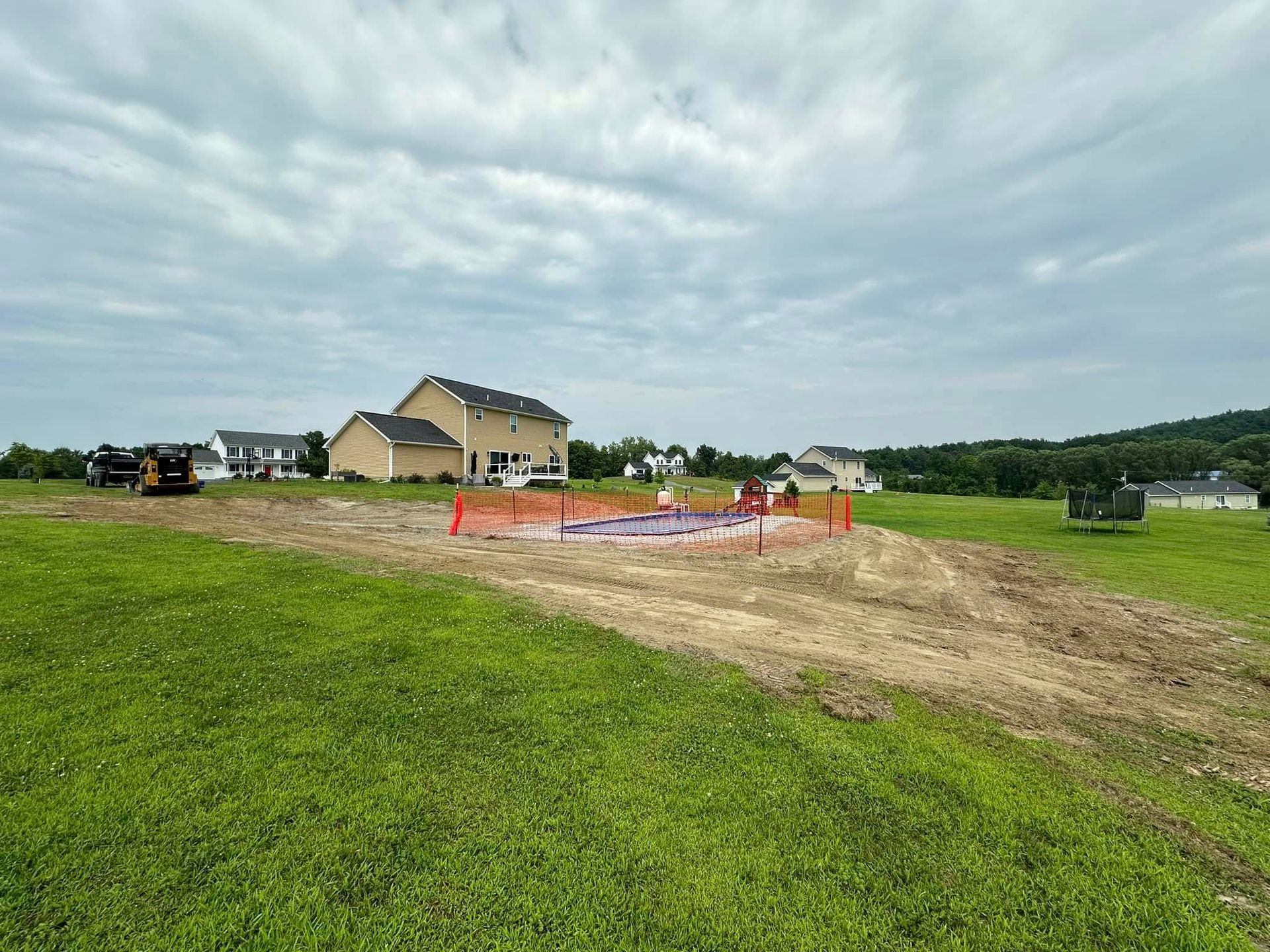 Construction site in field, with a house in the background and cloudy sky. Orange safety fence surrounds an area.