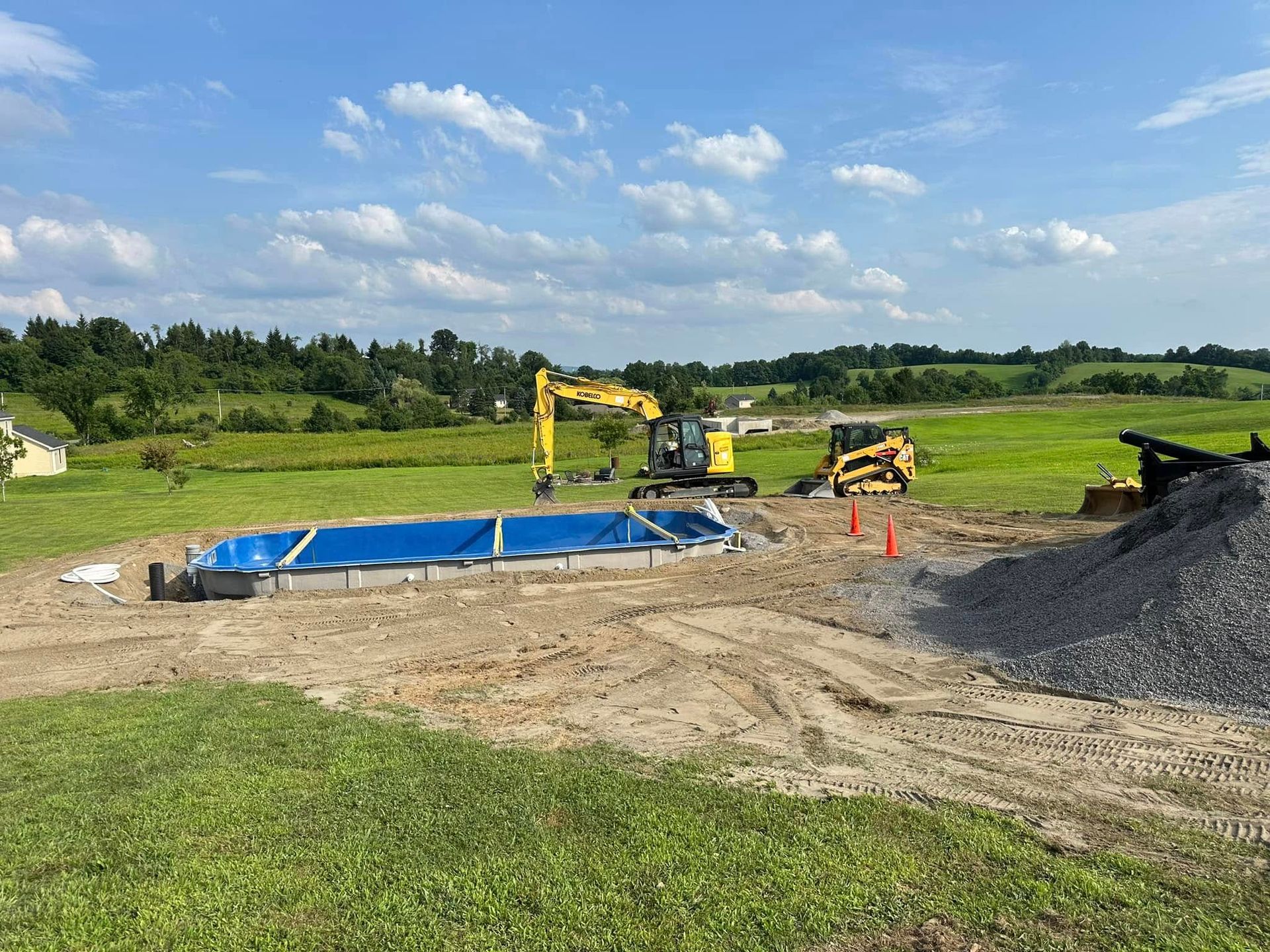 Construction site: excavator and equipment surrounding a rectangular pool with blue tarp. Sunny day, open field.