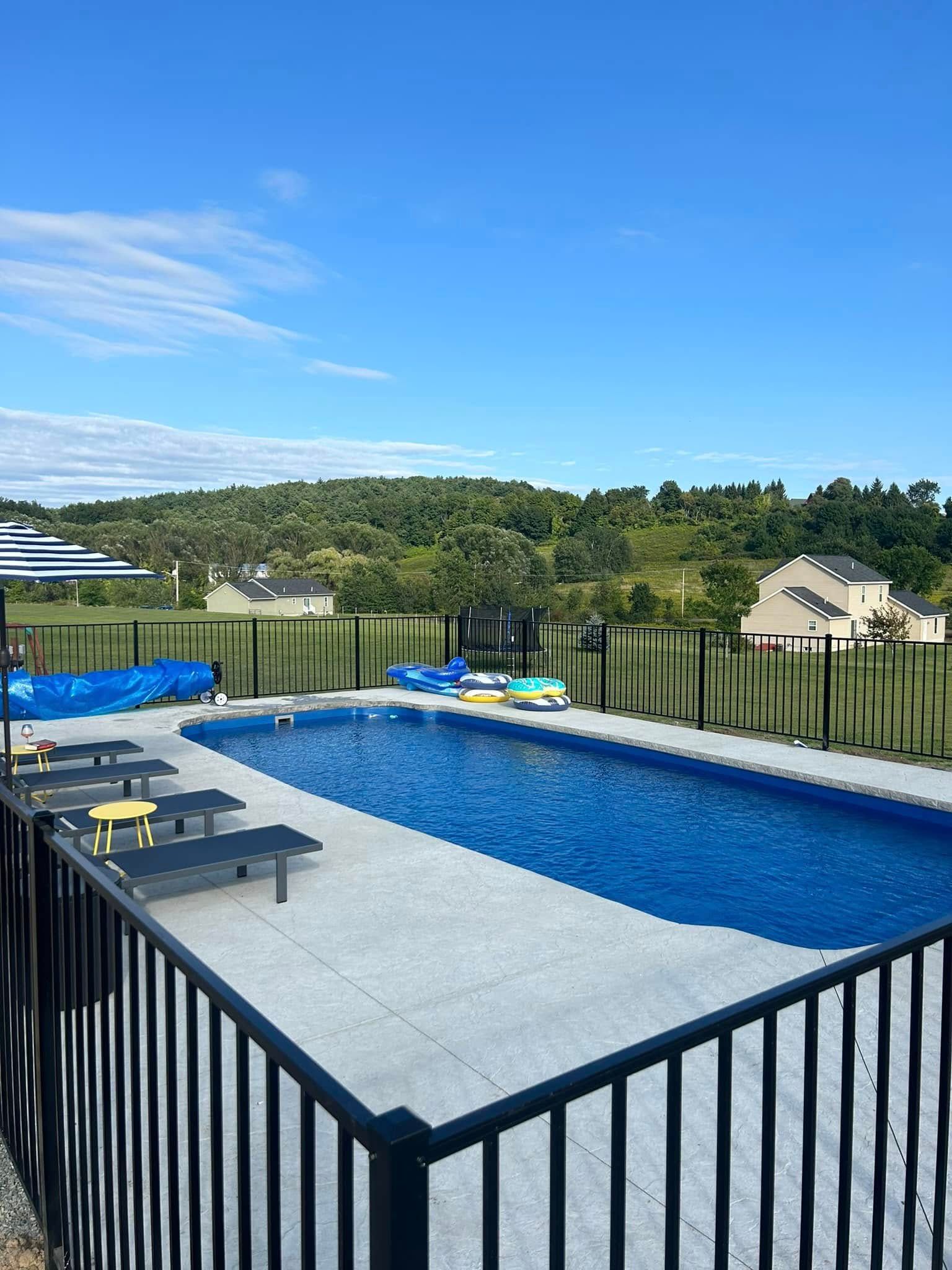 A rectangular pool with blue water sits in a yard with a black fence, blue sky, and green hills in the background.