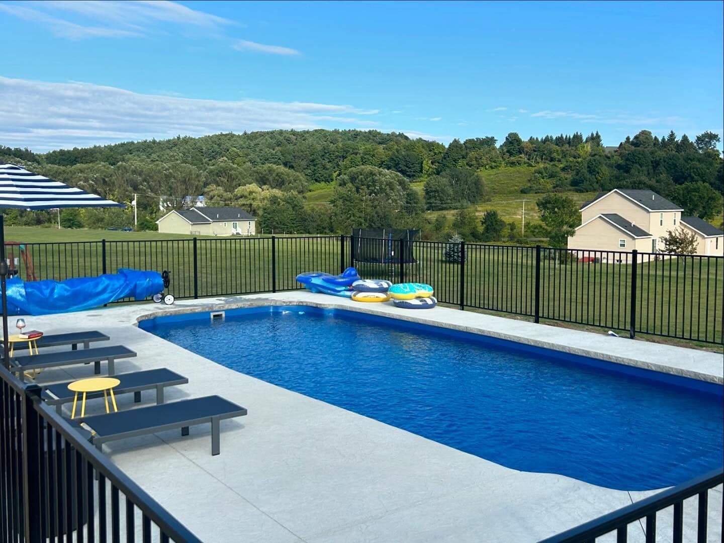 Pool with blue water and surrounding patio; fenced yard with houses and hills in the background.