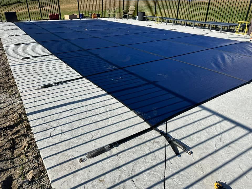 A rectangular pool covered with a dark blue safety cover, surrounded by concrete and a black fence.