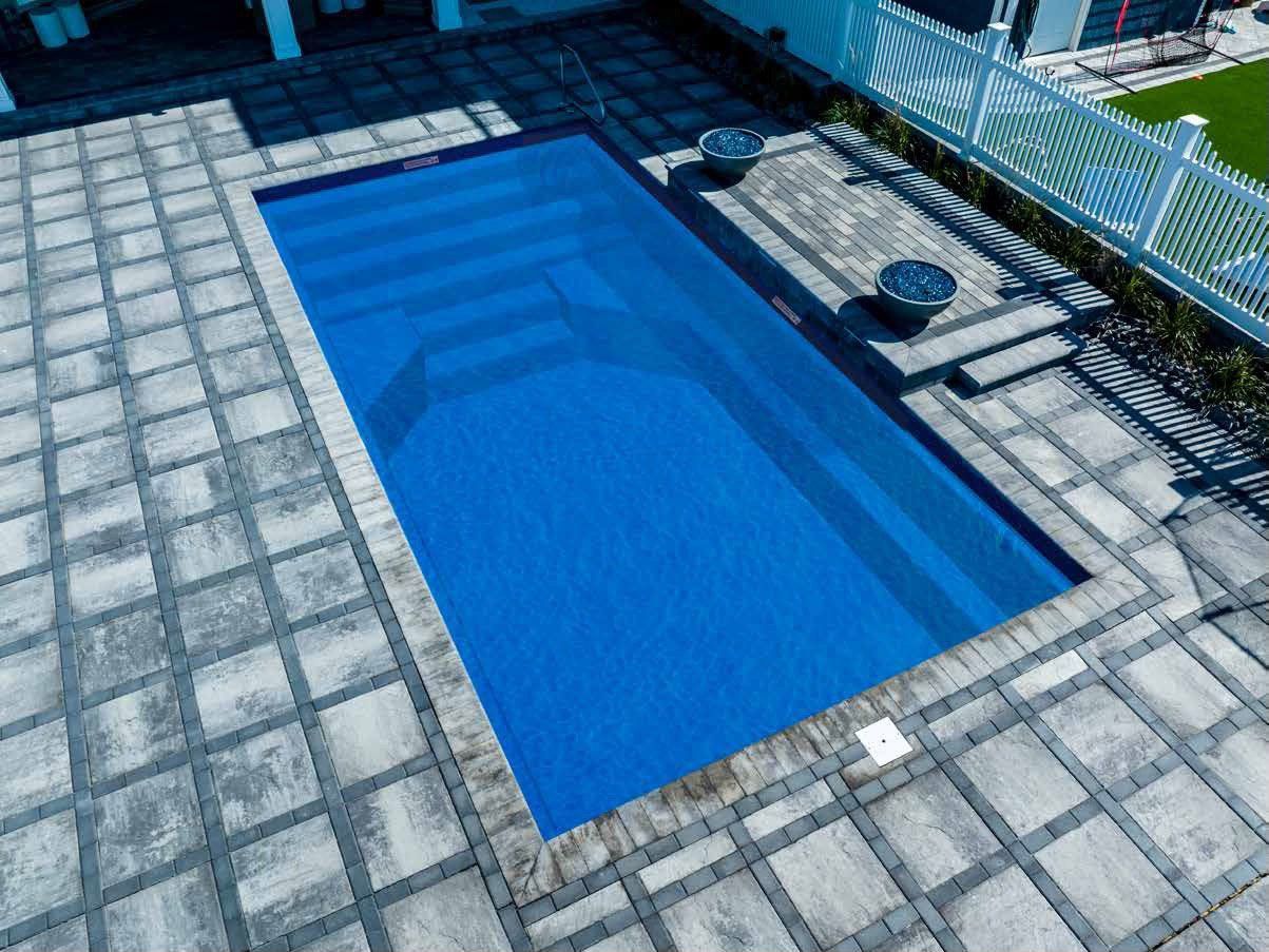 Rectangular in-ground swimming pool with blue water, surrounded by grey pavers and steps, in an outdoor setting.