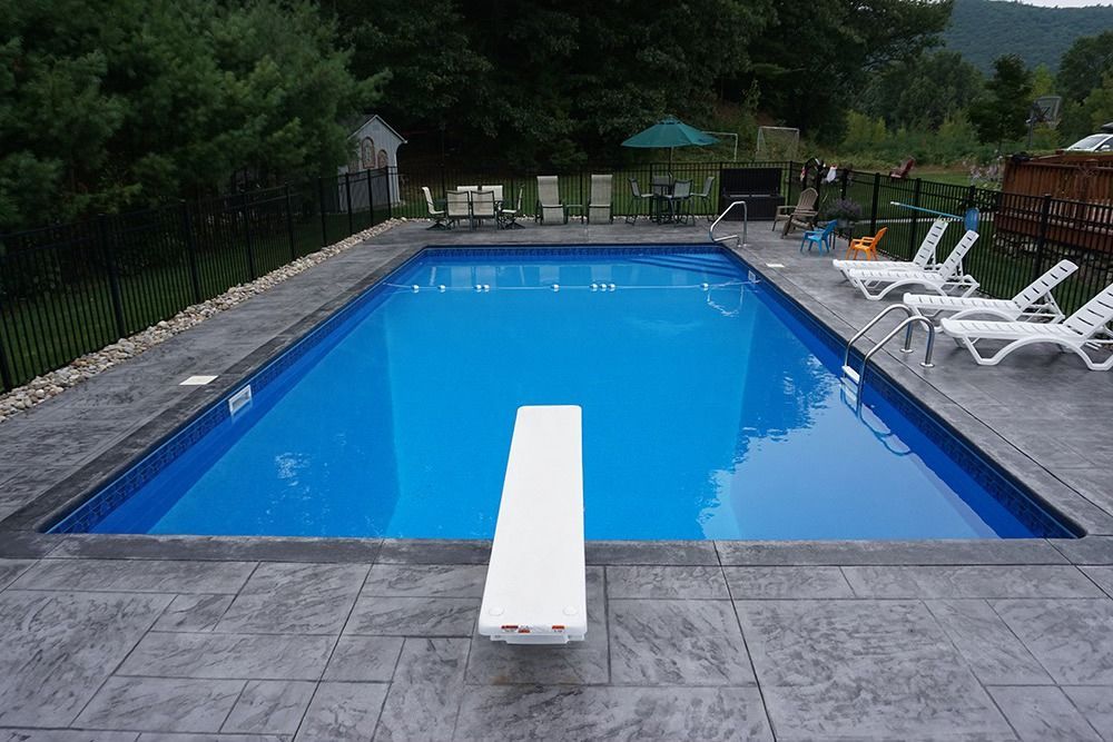Rectangular blue swimming pool with diving board, surrounded by gray concrete patio.