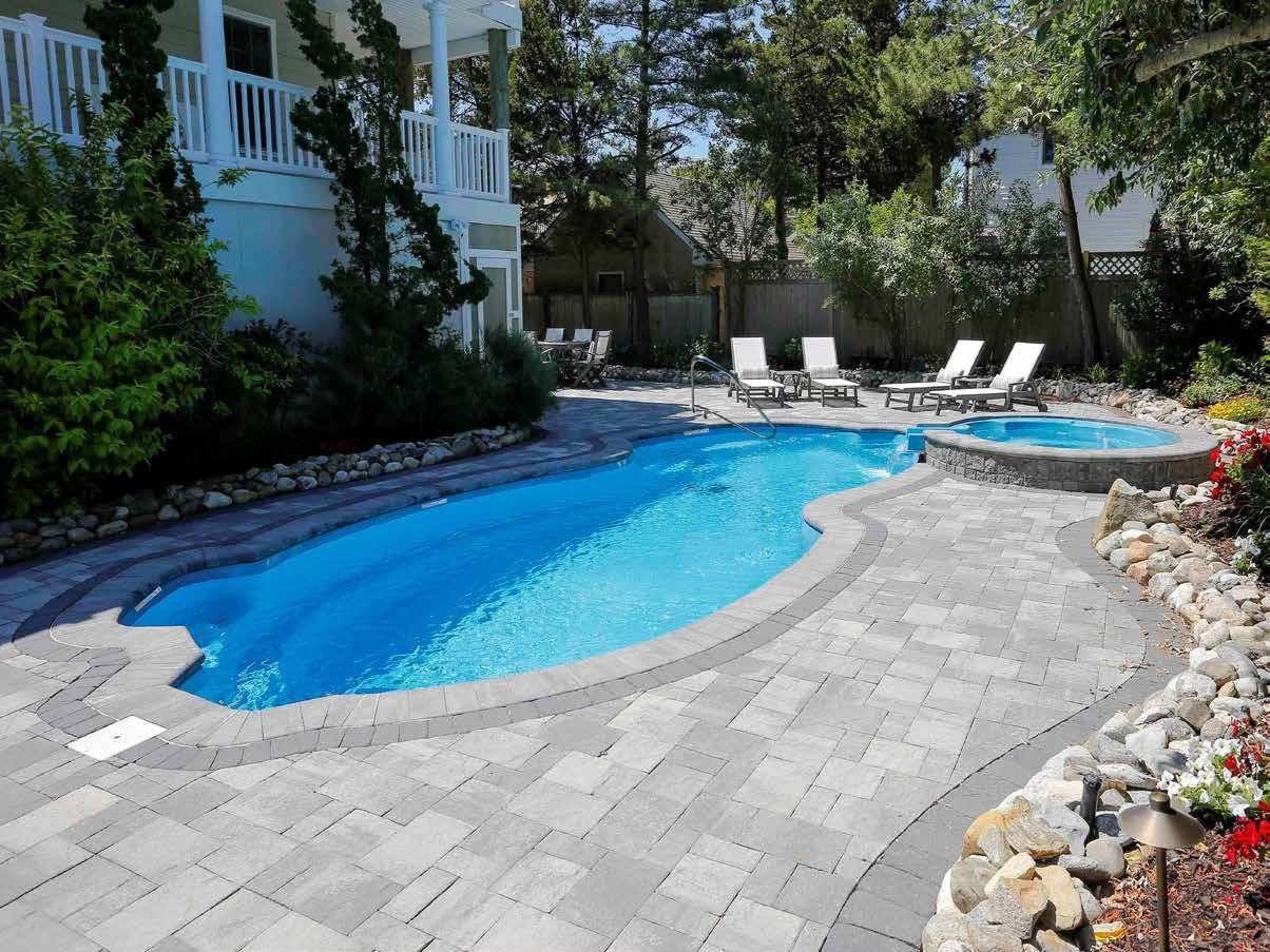 A backyard pool with blue water and a hot tub, surrounded by grey pavers, lounge chairs, and landscaping.