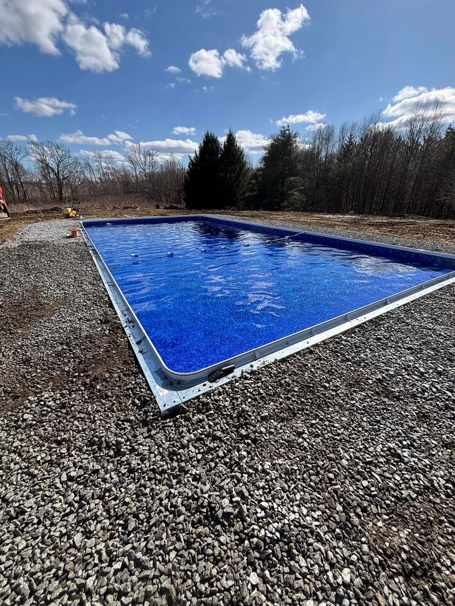 Rectangular blue pool surrounded by gravel against a backdrop of trees and a blue sky with clouds.