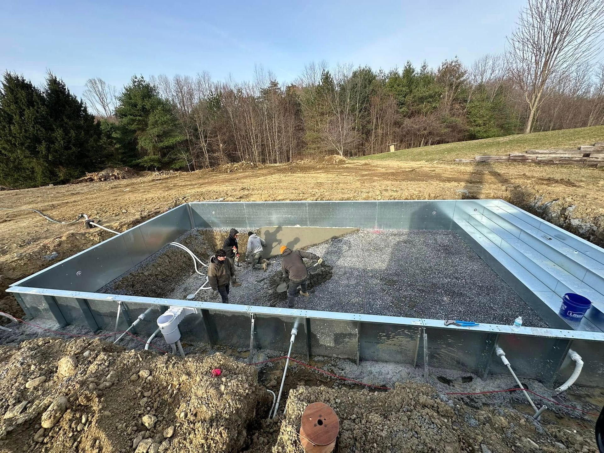Pool under construction in a dirt yard, with gravel inside. People working, trees in background, sunny day.