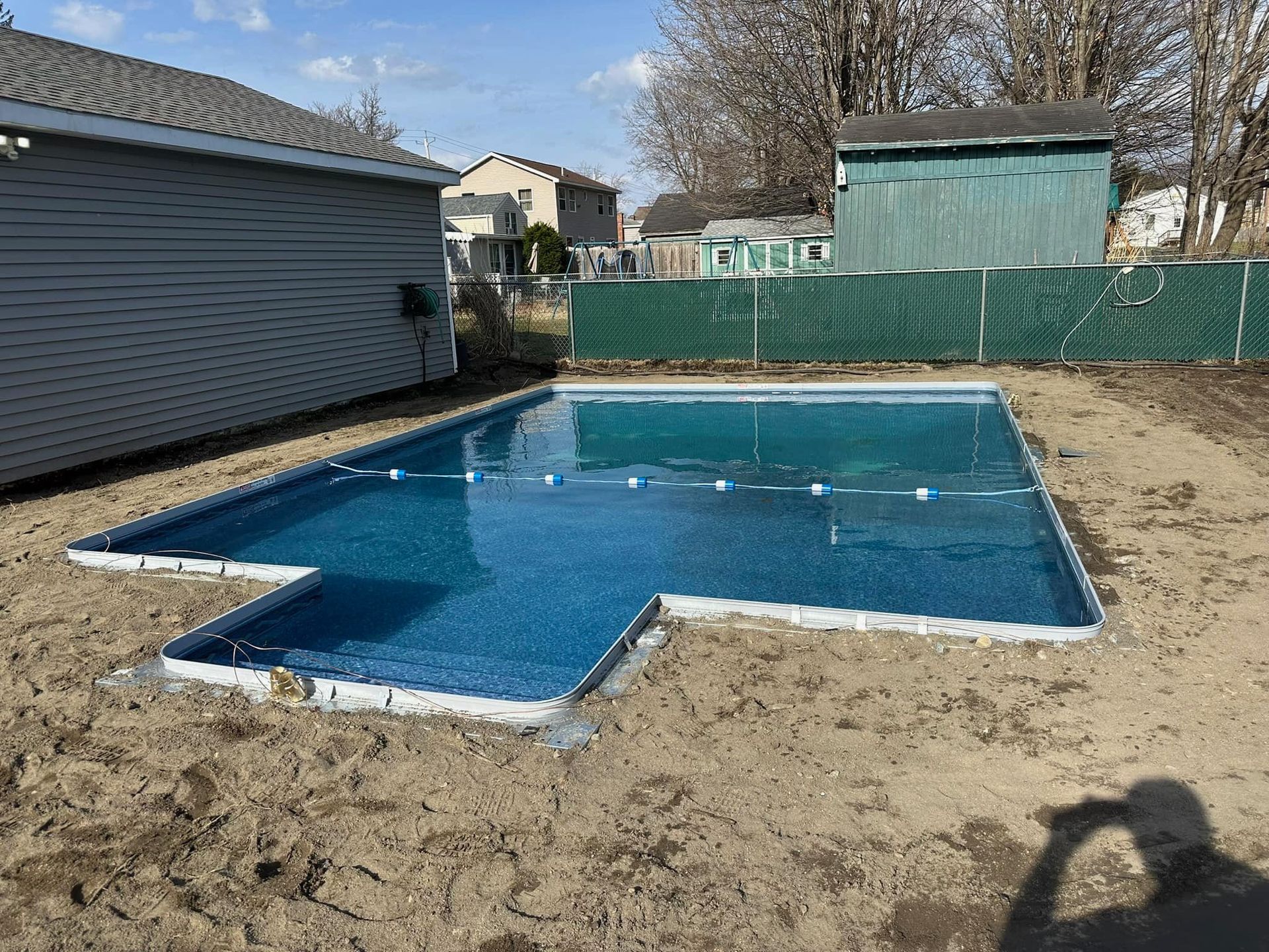 Rectangular in-ground swimming pool with blue water. Pool is surrounded by dirt and concrete. Houses and fence in background.