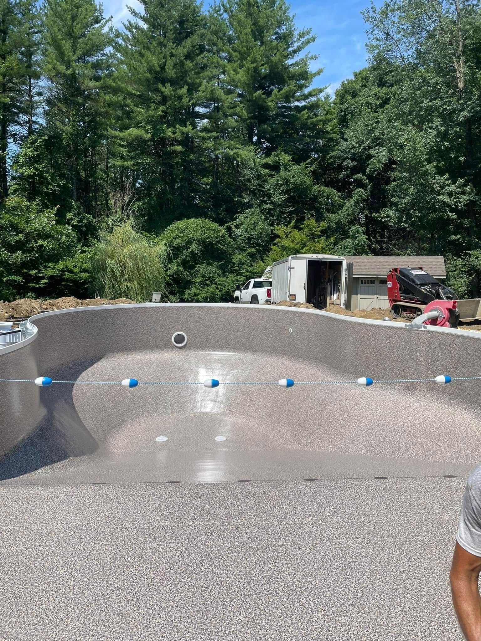 Pool under construction, with dark-speckled interior, blue and white pipes, surrounded by trees.