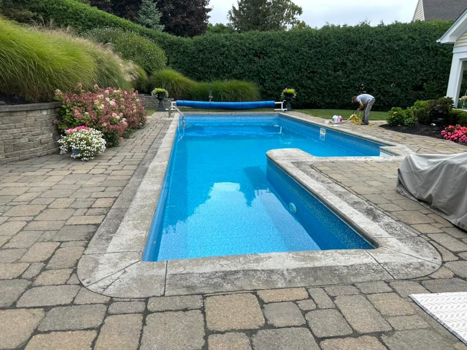 A rectangular swimming pool surrounded by stone pavers, shrubs, and a person working nearby.