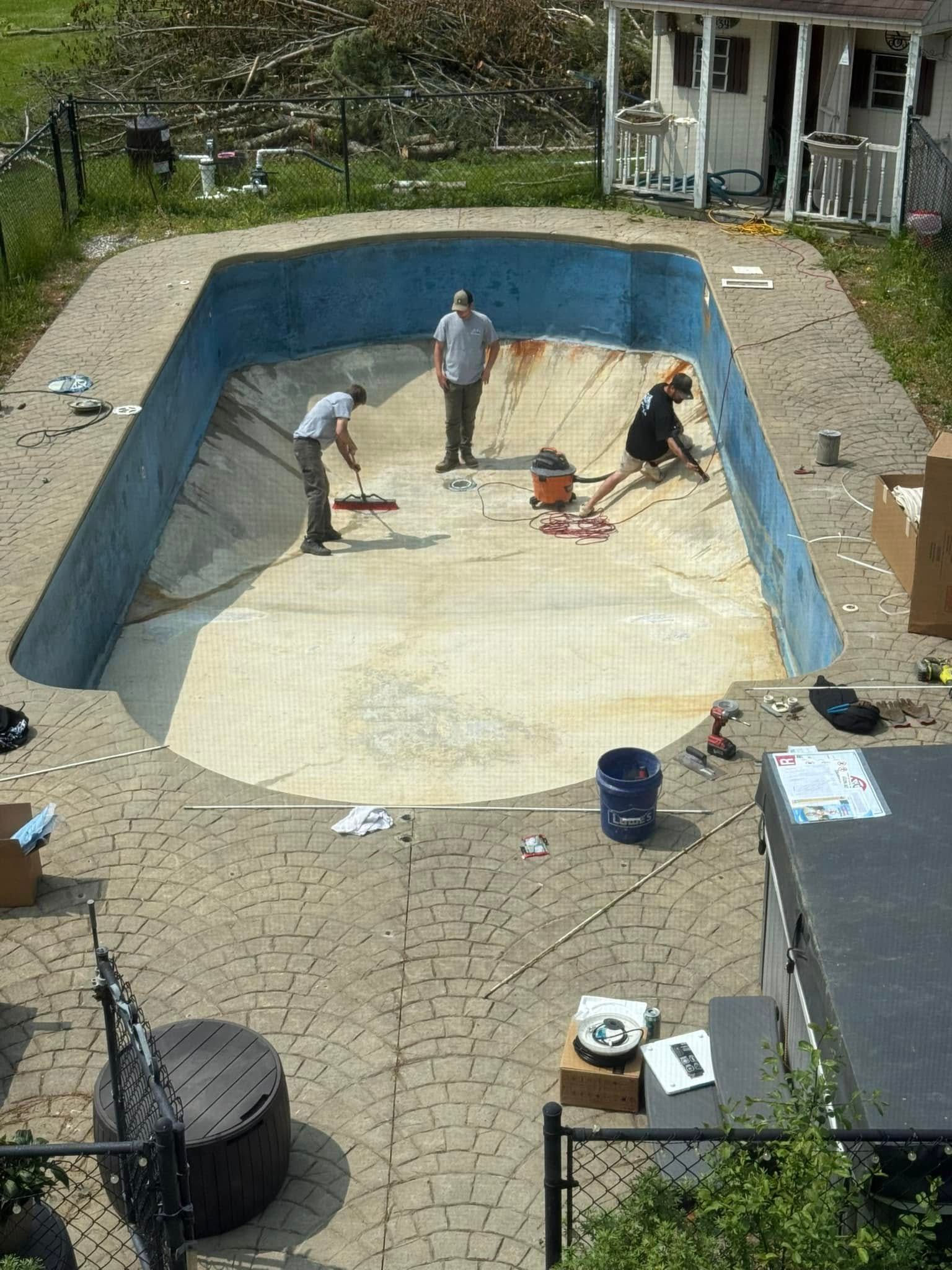 Workers cleaning an empty, rectangular swimming pool with blue painted walls and paved surround.
