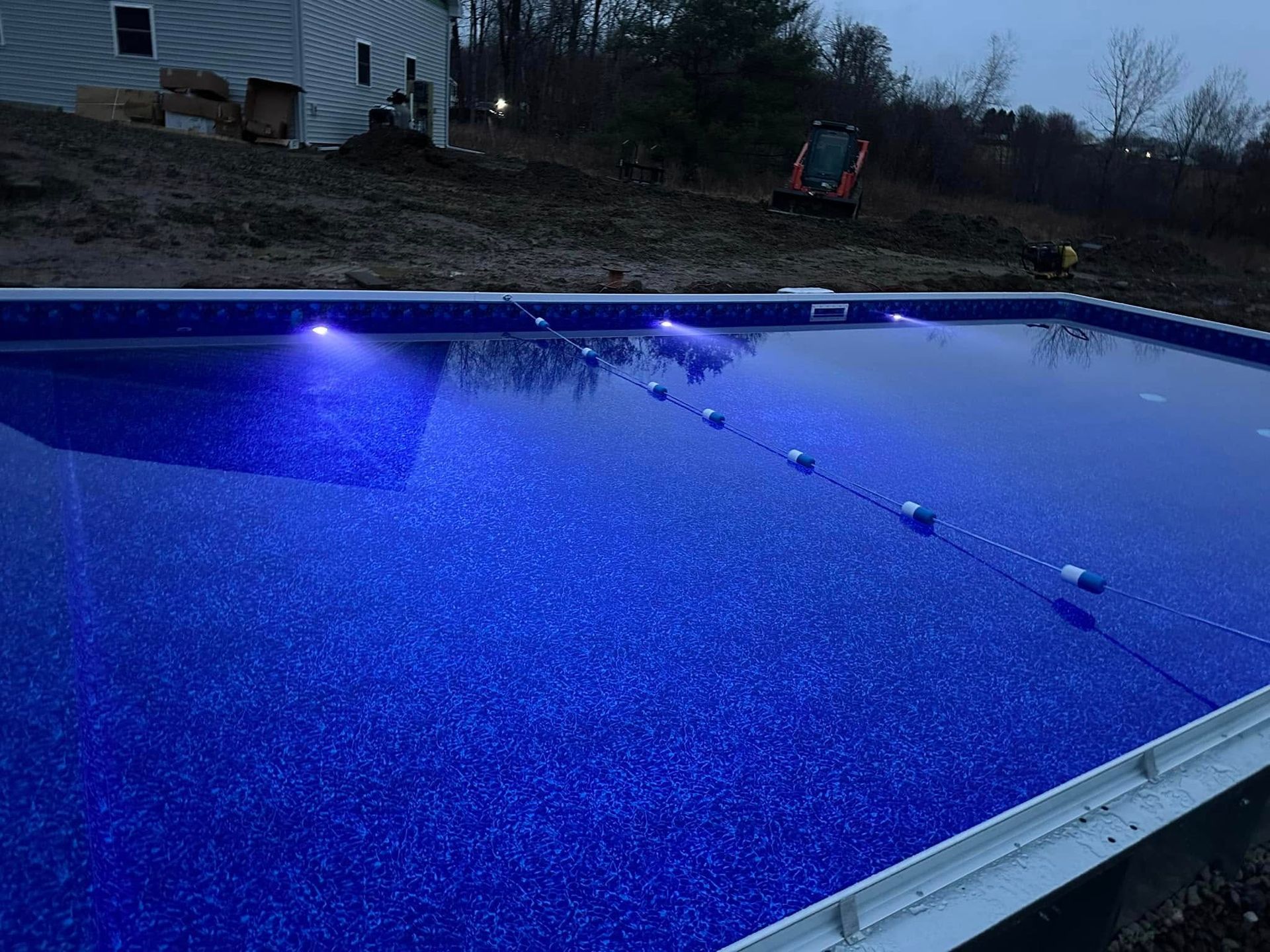 Blue-lit rectangular pool at dusk; construction site background; white coping; string of buoys.