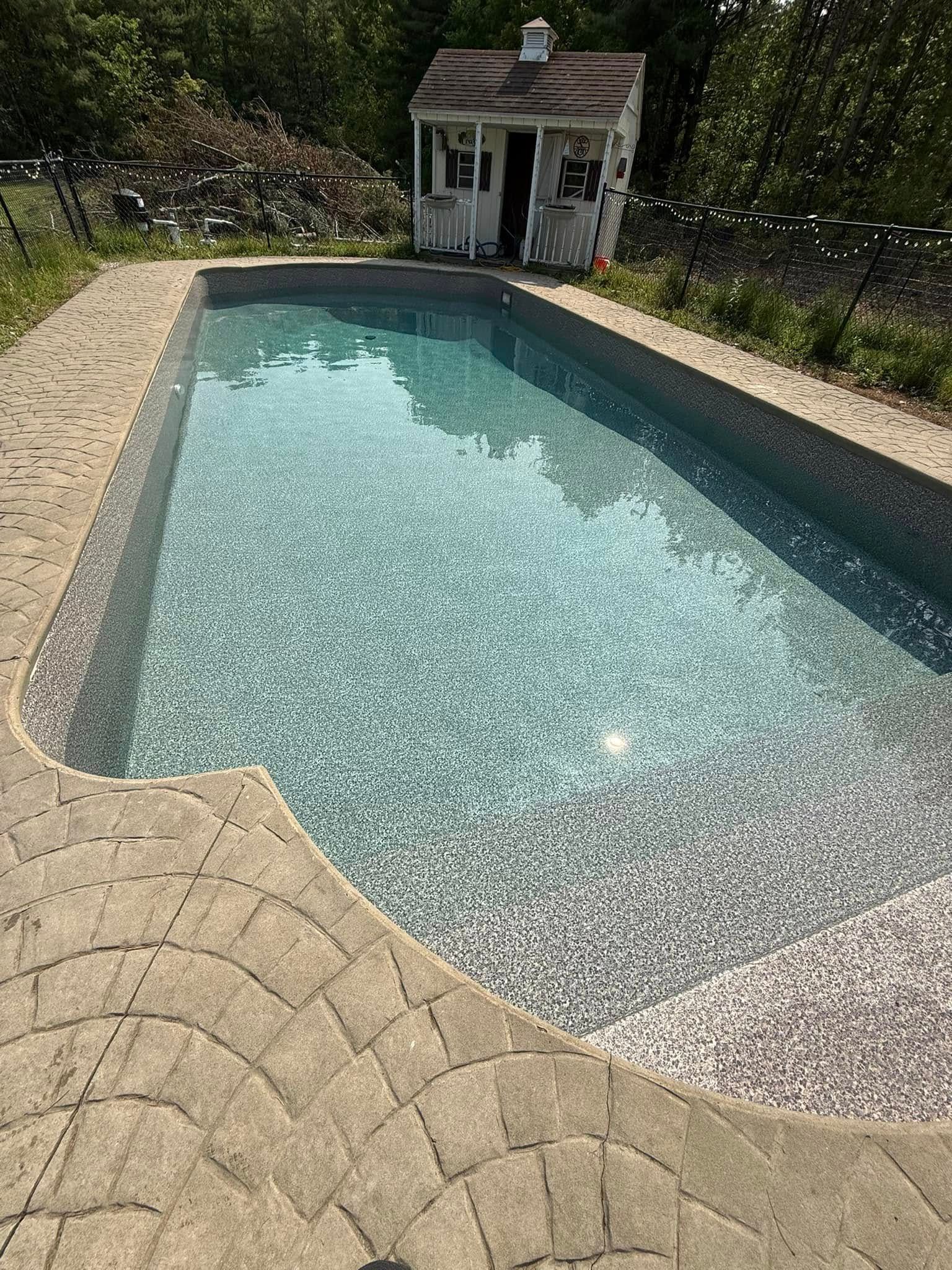 Rectangular swimming pool with blue water, surrounded by stone paving and a small white shed in the background.