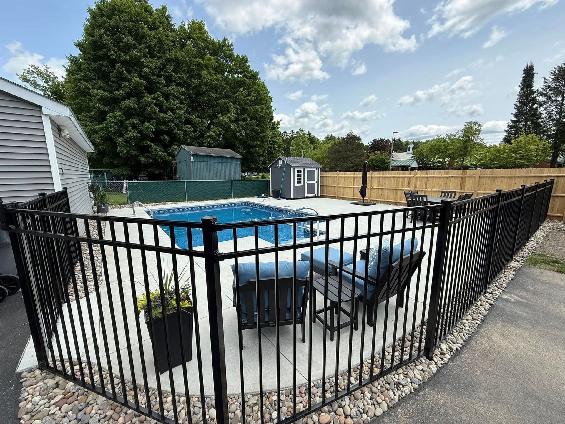 Black fenced-in backyard pool area with lounge chairs on concrete patio and small sheds beyond.