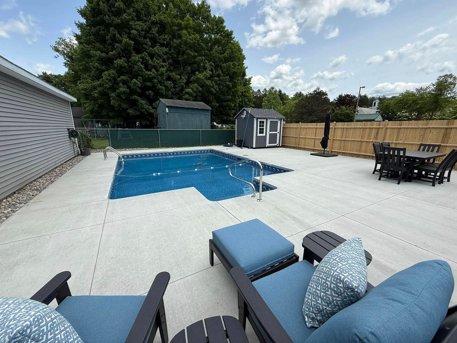Backyard with pool, lounge chairs, dining set, and sheds, surrounded by a fence, on a sunny day.